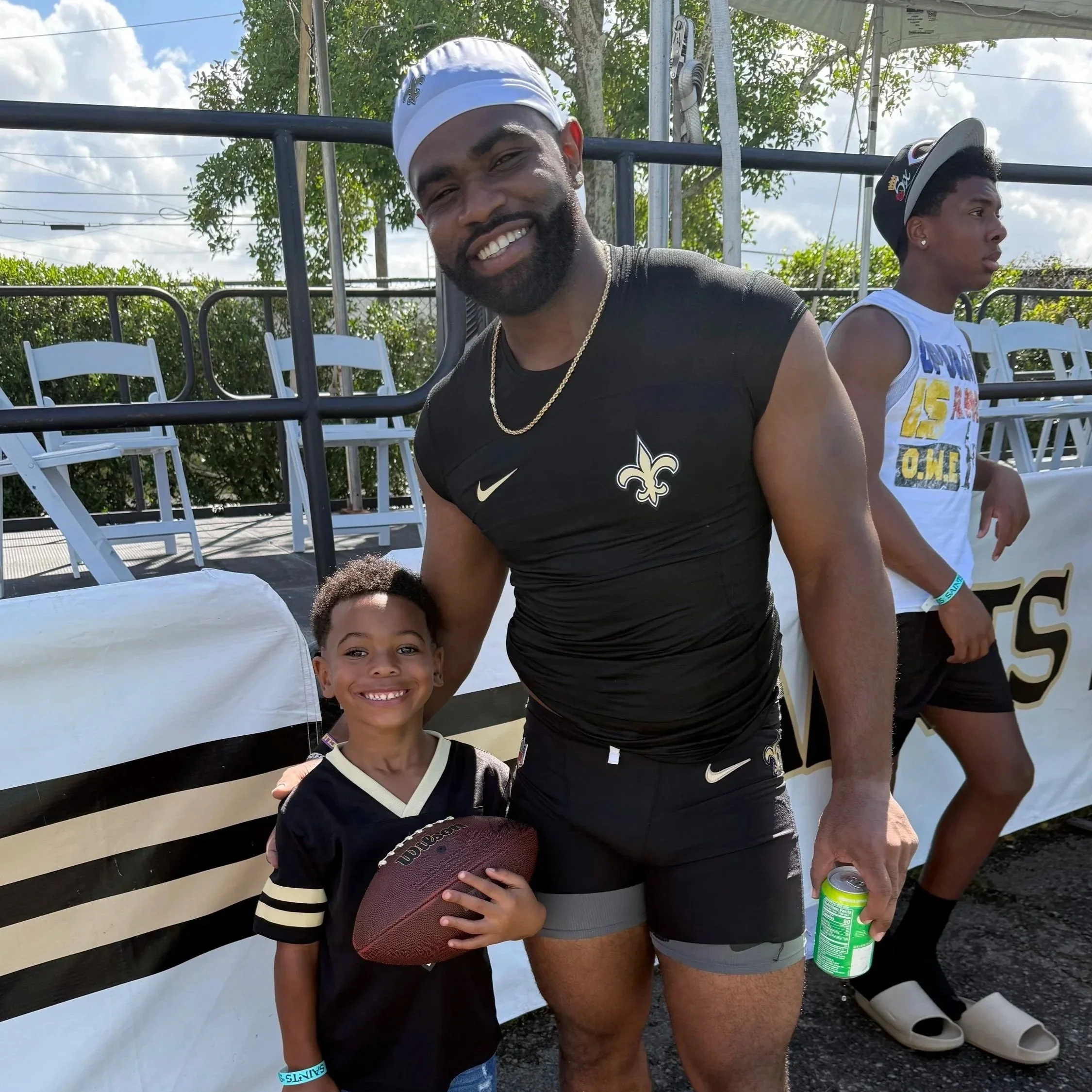 A smiling man in a New Orleans Saints black sports uniform, wearing a white cap and gold chain, standing next to a young boy in a black football jersey holding a football, outdoors with white chairs and people in the background.