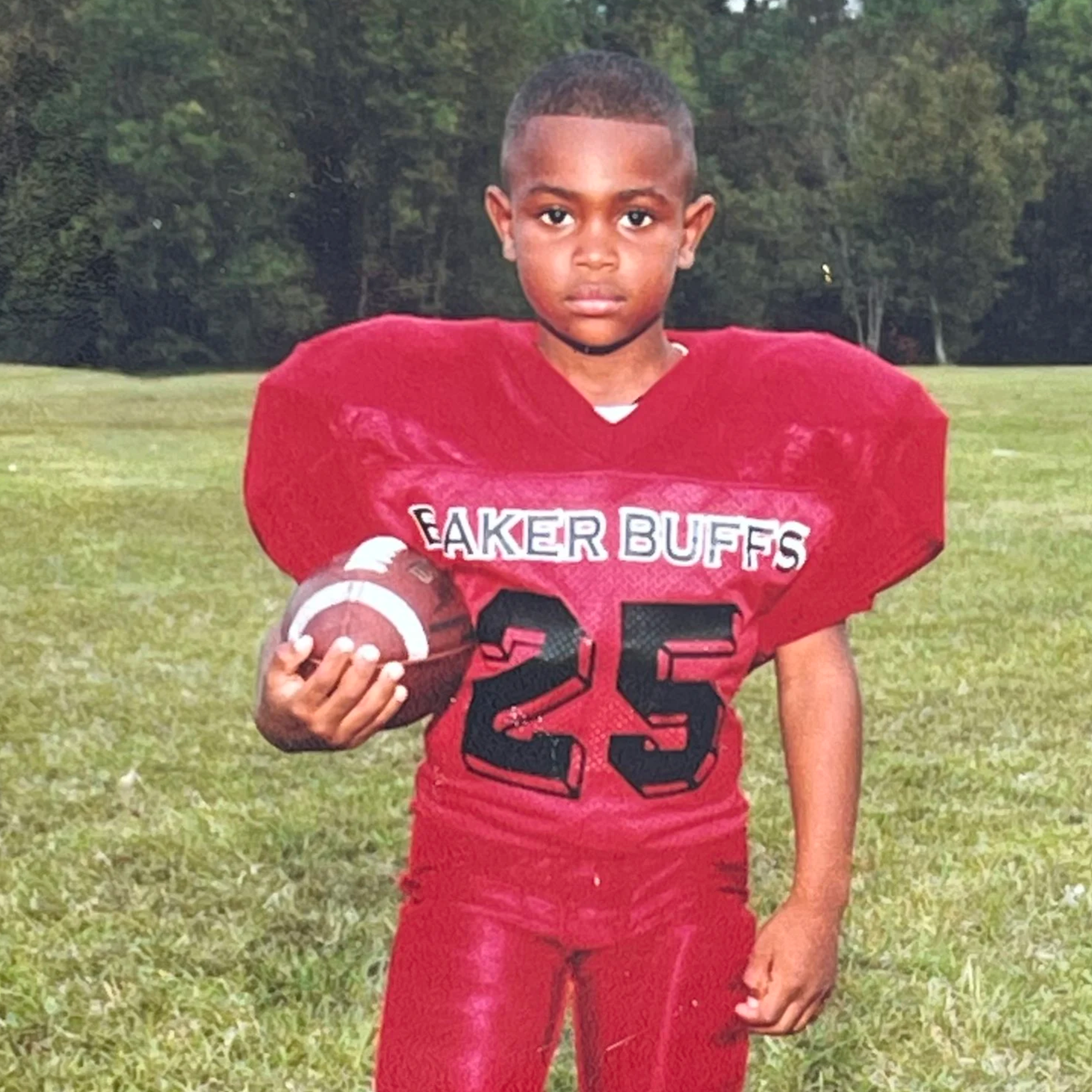 A young boy in a red football uniform holding a football, standing on a grassy field with trees in the background.