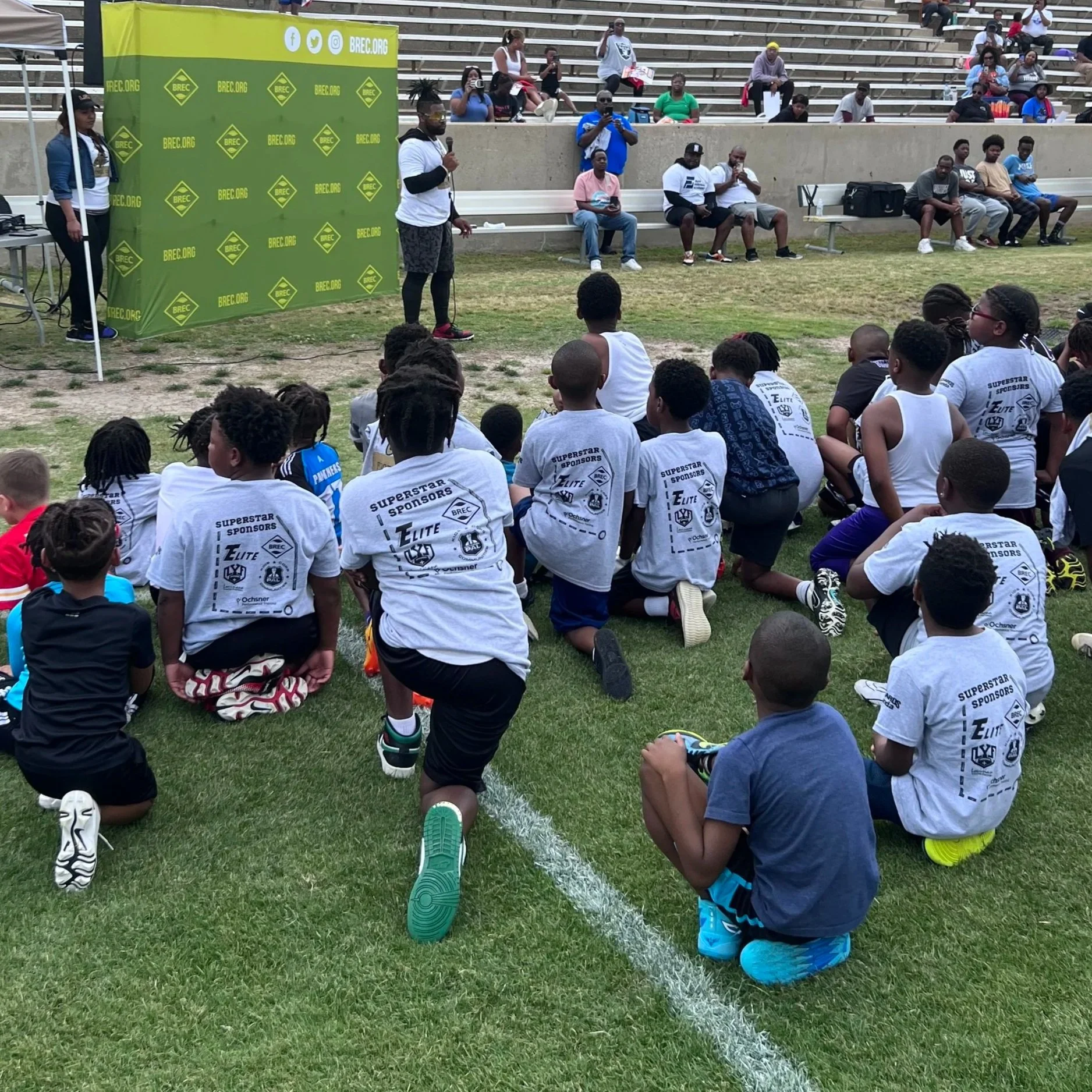 A group of young children sitting and kneeling on a grassy field, attending an outdoor event with a speaker and audience seated on bleachers behind them. The event promotes BRECS.org.