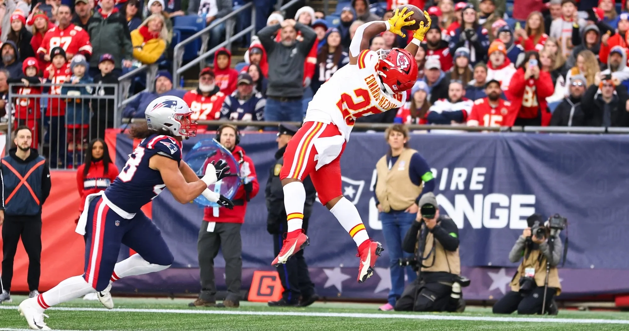 An American football player in Kansas City Chiefs uniform catches a football in the end zone as a New England Patriots player tries to defend, during a game with cheering fans in the background.