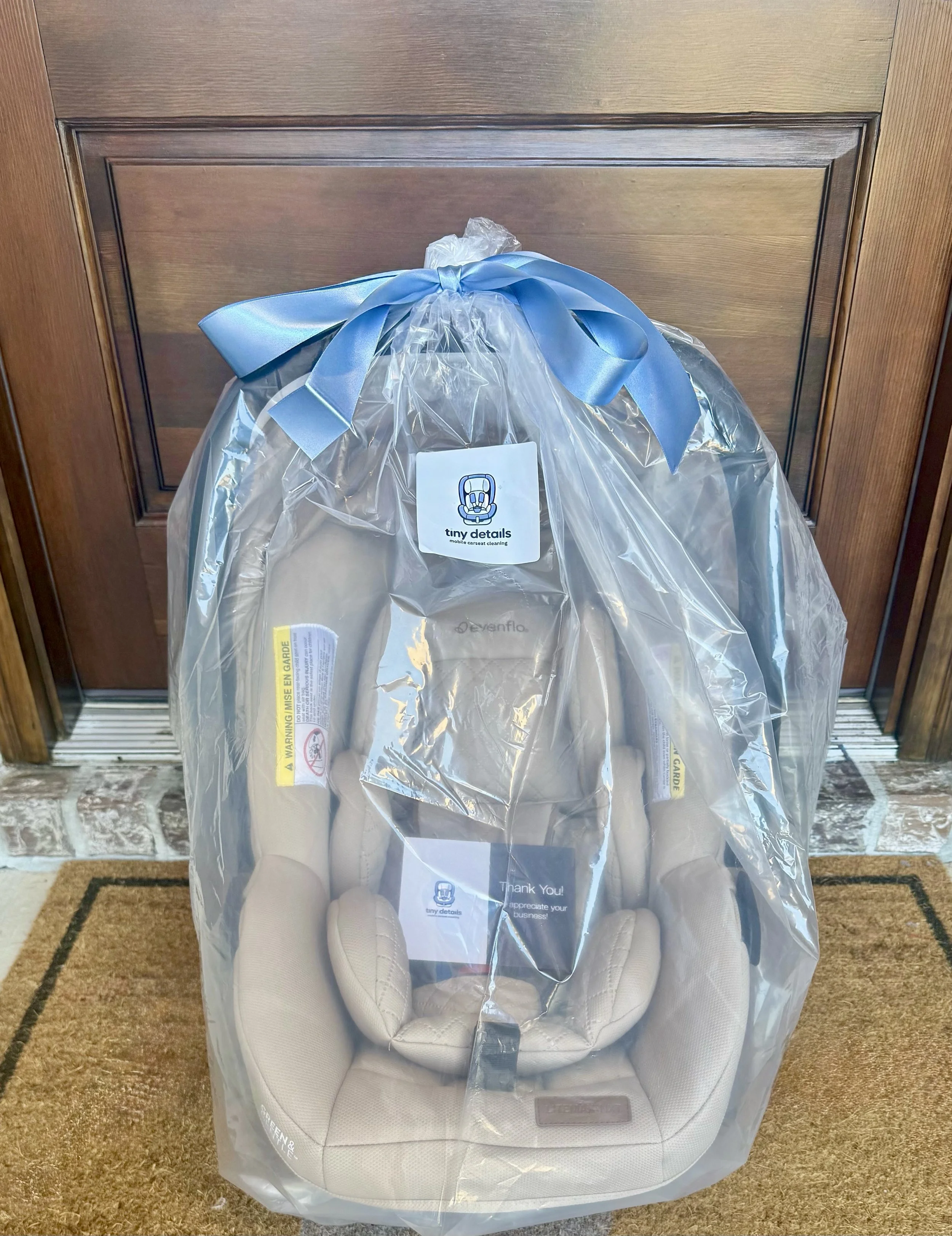 A baby car seat inside a clear plastic bag with a blue ribbon on top, placed in front of a wooden door.