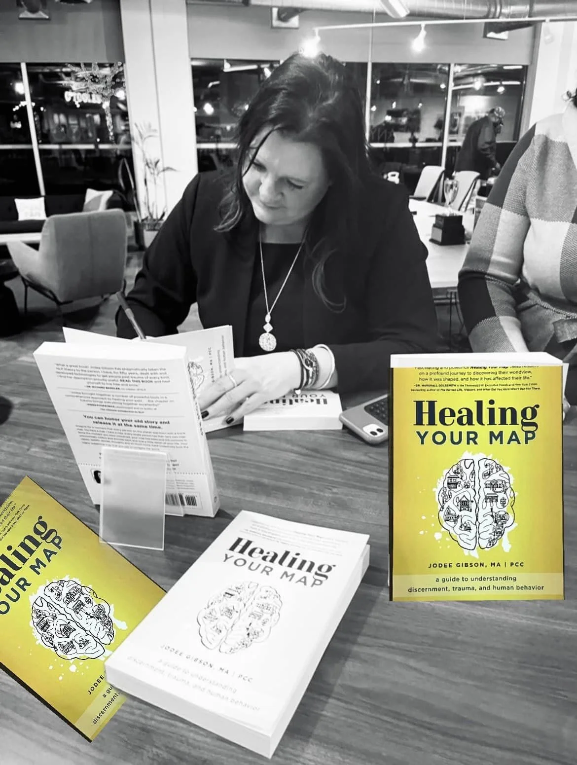A woman sitting at a table signing a book titled 'Healing Your Map,' with multiple copies of the book on the table. The book is yellow with black text and a brain illustration on the cover.