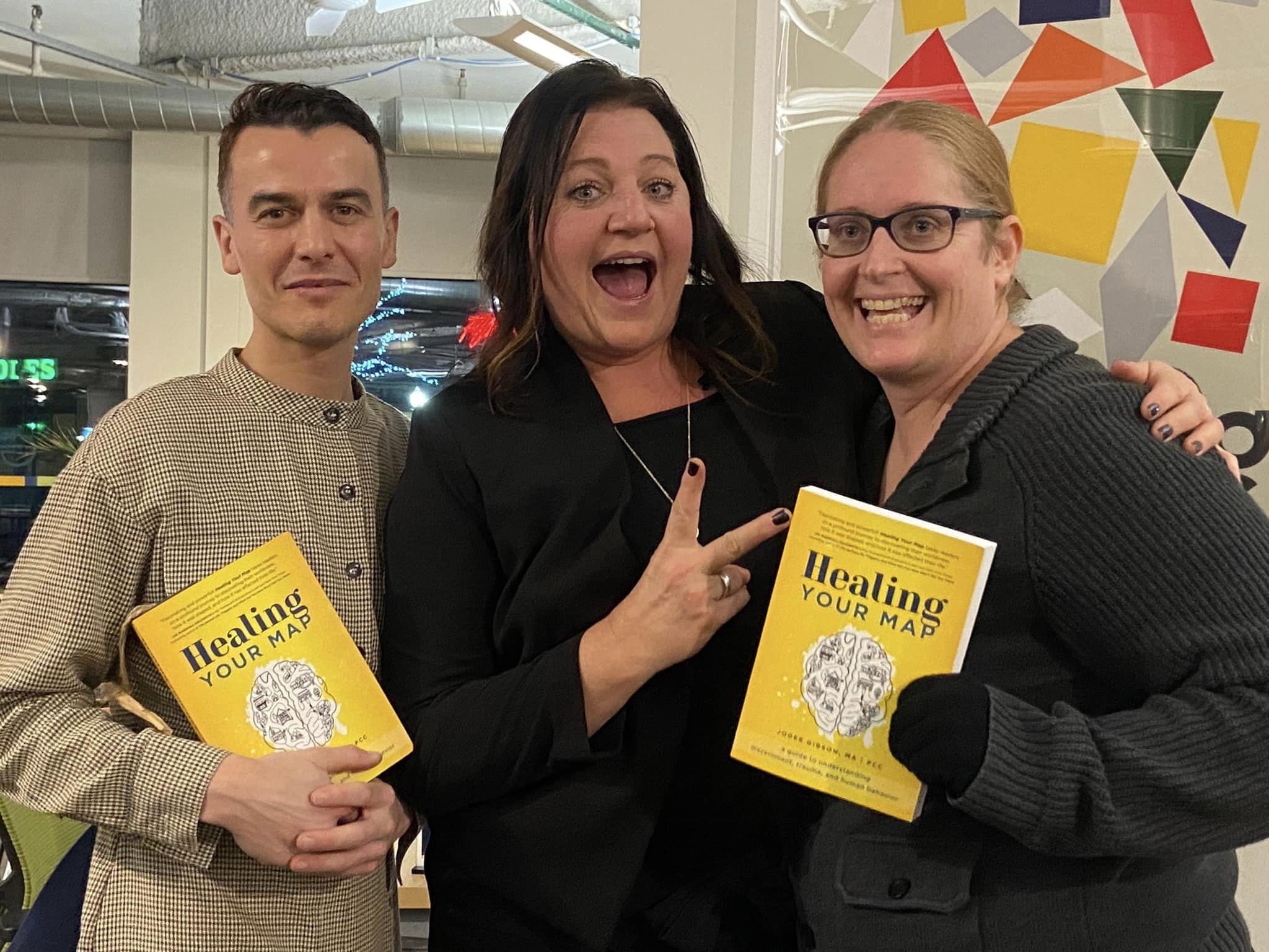 Three people smiling, Goran Mrvic, Jodee Gibson and Penny Roberts, holding books titled 'Healing Your Map,' in an indoor setting with colorful geometric wall art in the background.