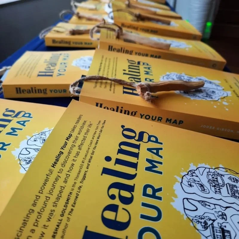 A row of yellow cards on a table, each with the title Healing Your Map and a wooden stick attached. The cards feature an illustration of a brain with various doodles related to healing and self-discovery.
