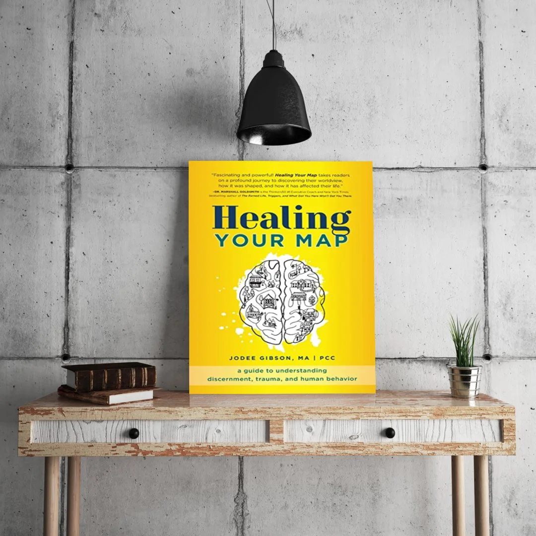 A yellow book titled "Healing Your Map" is displayed on a rustic wooden table beneath a black pendant lamp. The cover features an illustration of a brain with various buildings and objects inside. The background includes an industrial-style concrete 