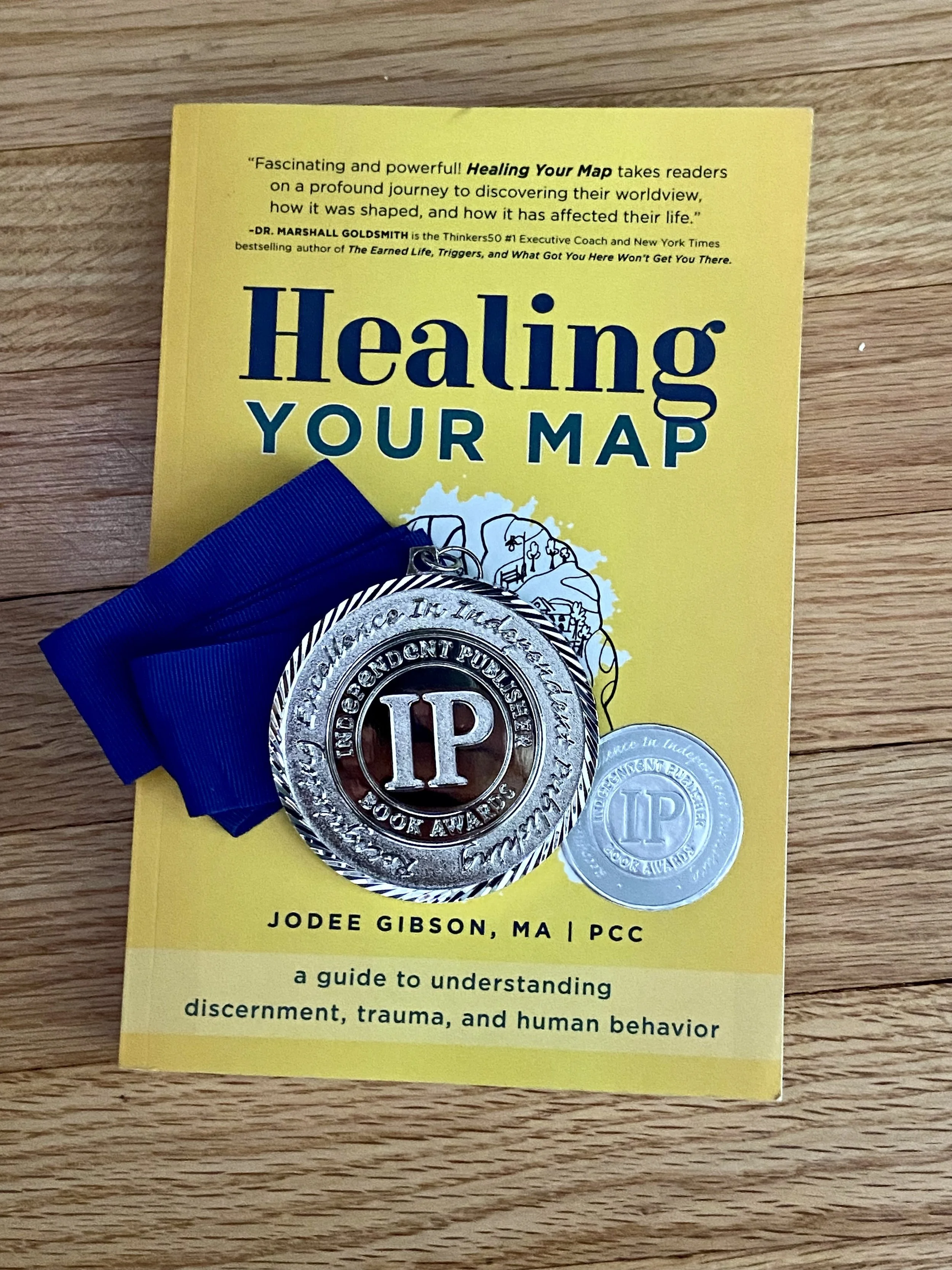 A yellow book titled 'Healing Your Map' by Jodee Gibson, MA, PCC, placed on a wooden surface, with a silver medal with blue ribbon on top of it.