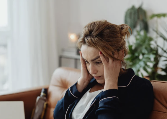 A woman sitting on a sofa holding her head with both hands, looking stressed or worried in a cozy living room.