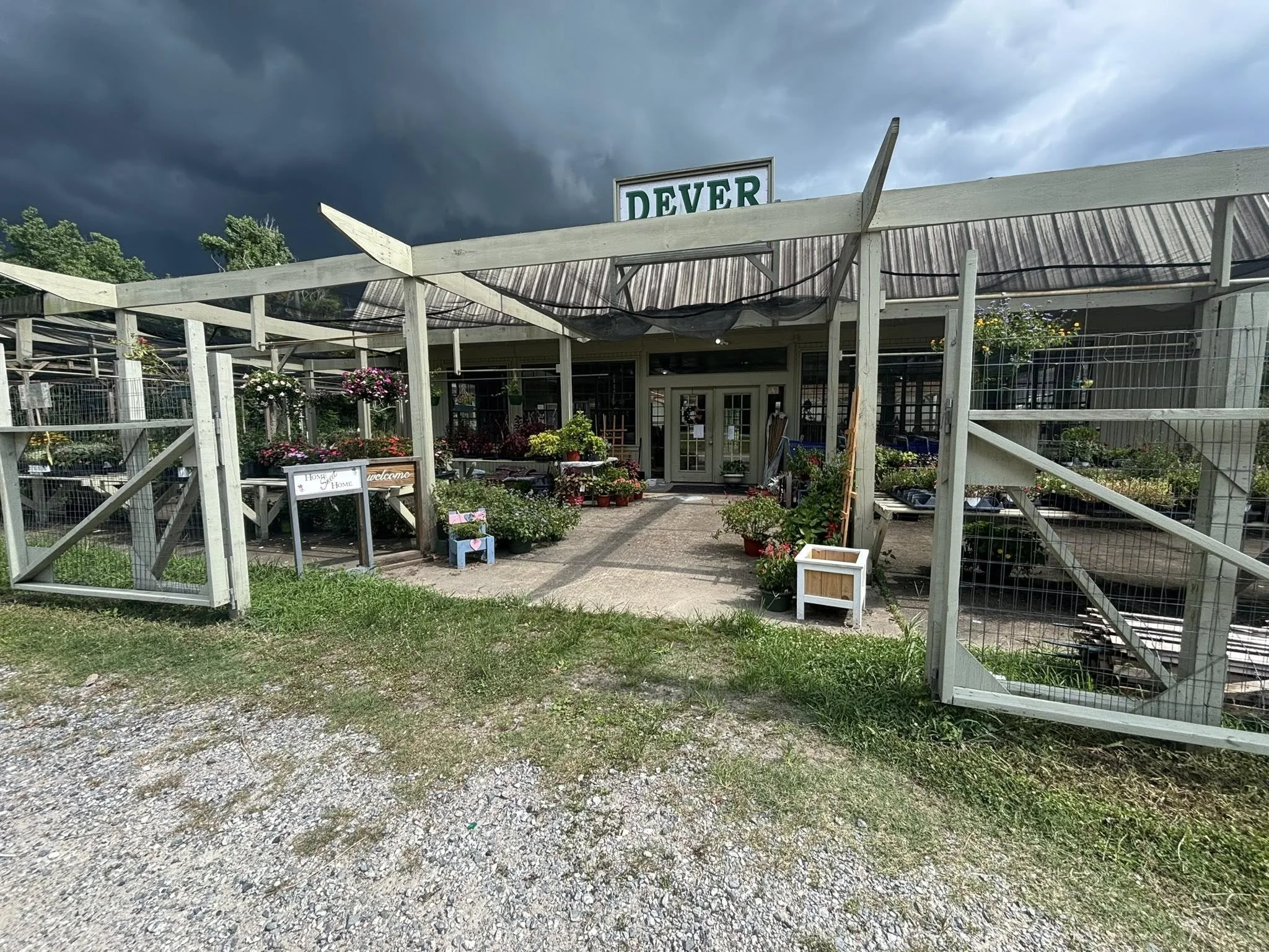 Exterior view of a garden shop and Plant Nursery at Dever Nursery in Southwest louisiana The entrance is decorated with potted plants and flowers, with a cloudy sky overhead.