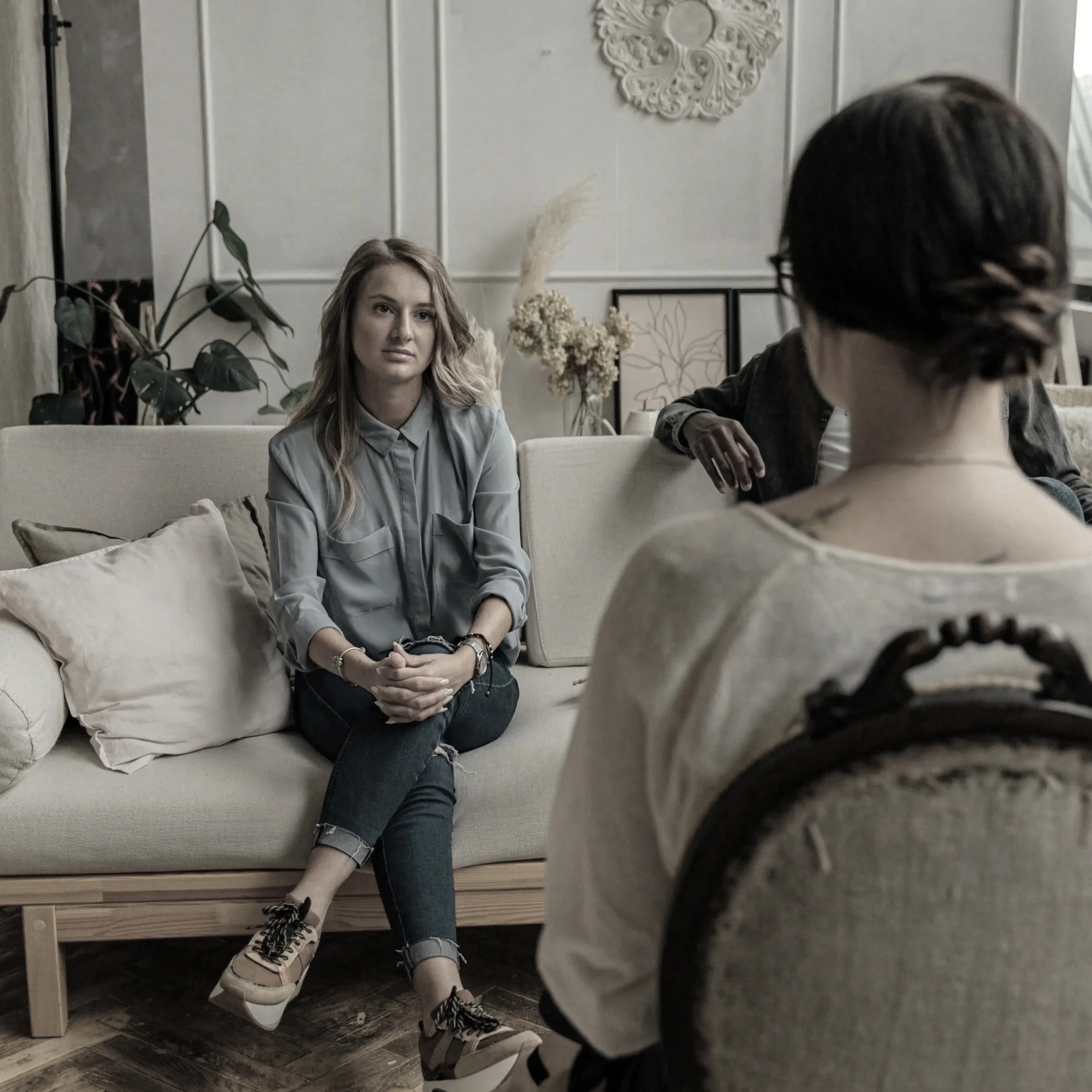A woman sitting on a beige sofa during a therapy session, talking to a therapist whose back is facing the camera.