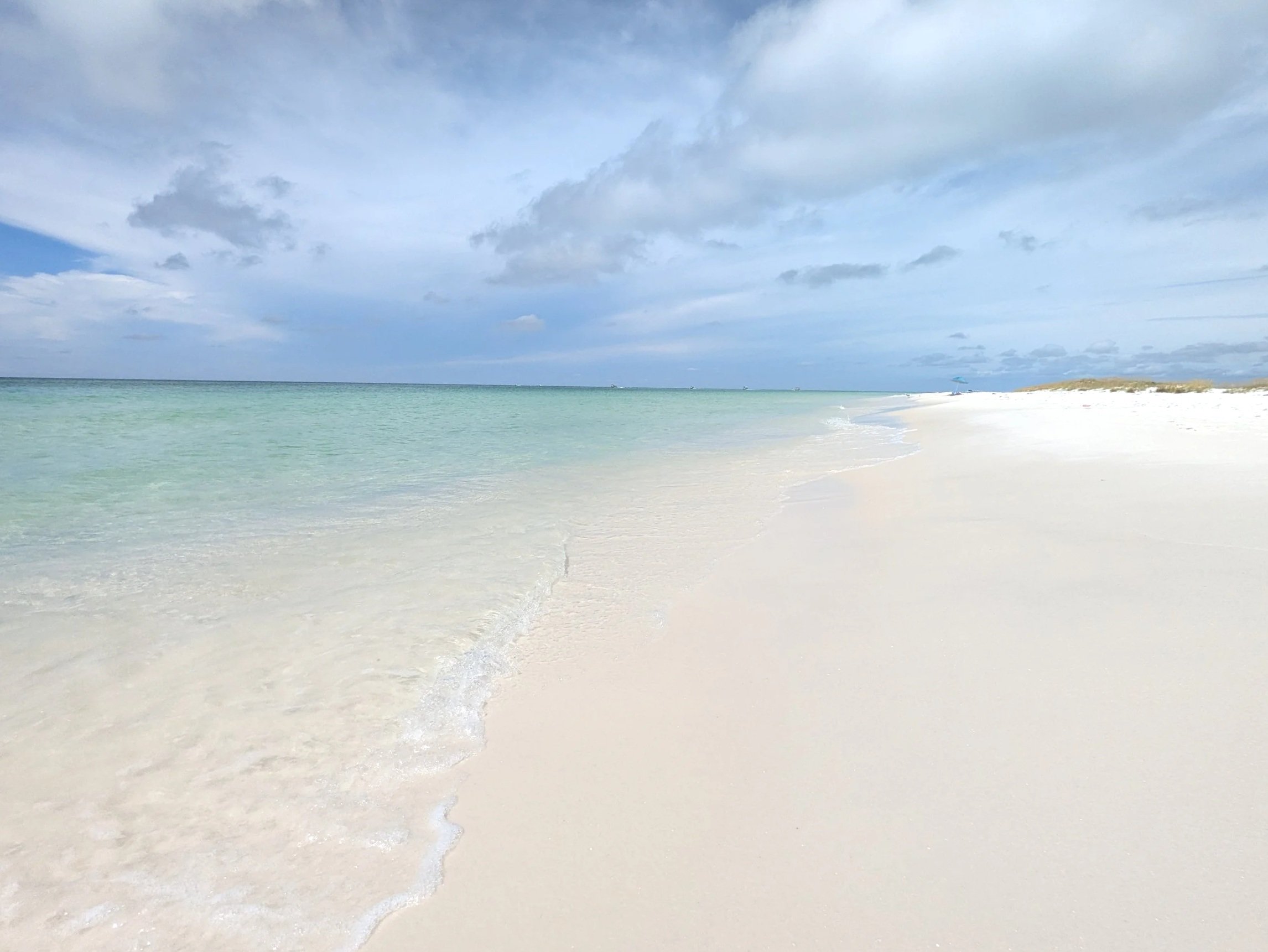 Empty white sandy beach with light turquoise water and a cloudy sky.