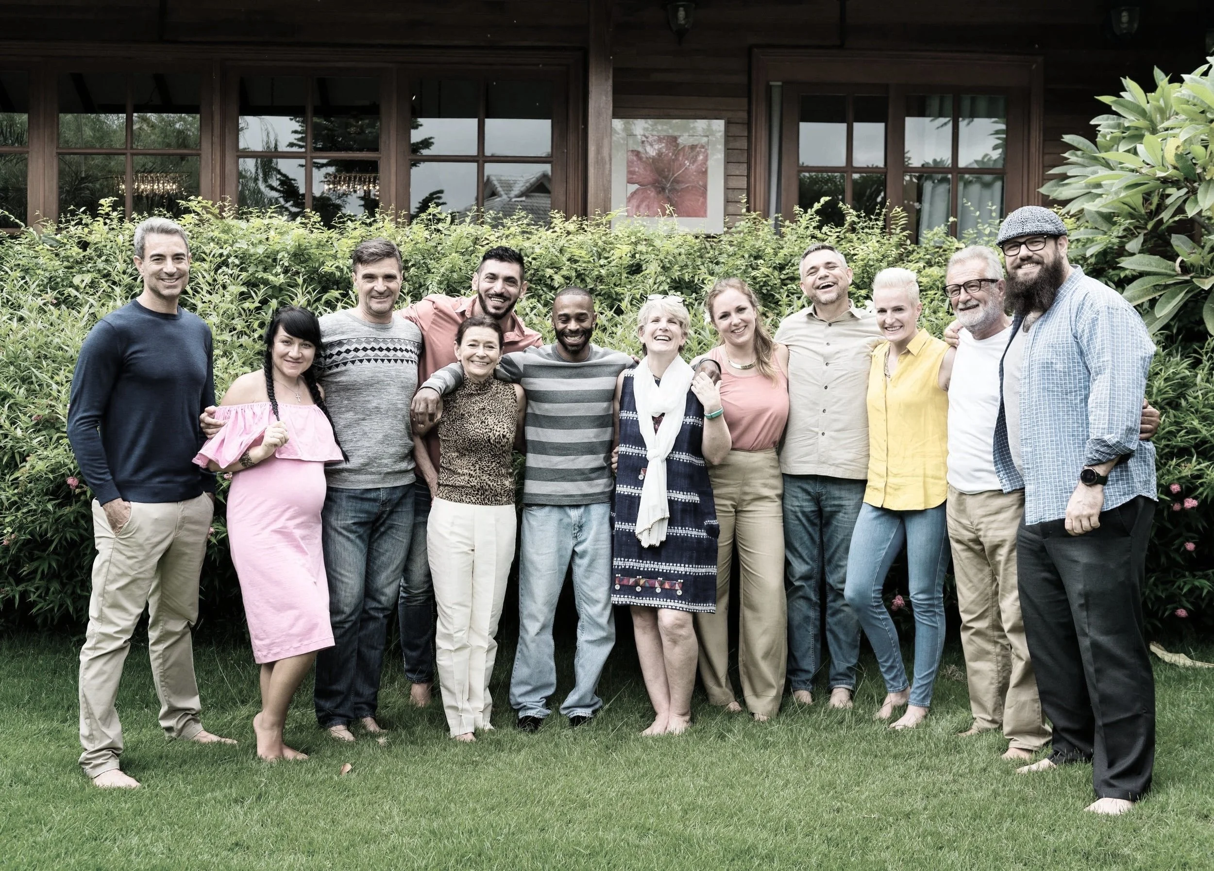 A diverse group of 14 people posing outside in front of a house with large windows and greenery. They are smiling and standing close together, with some having their arms around each other.