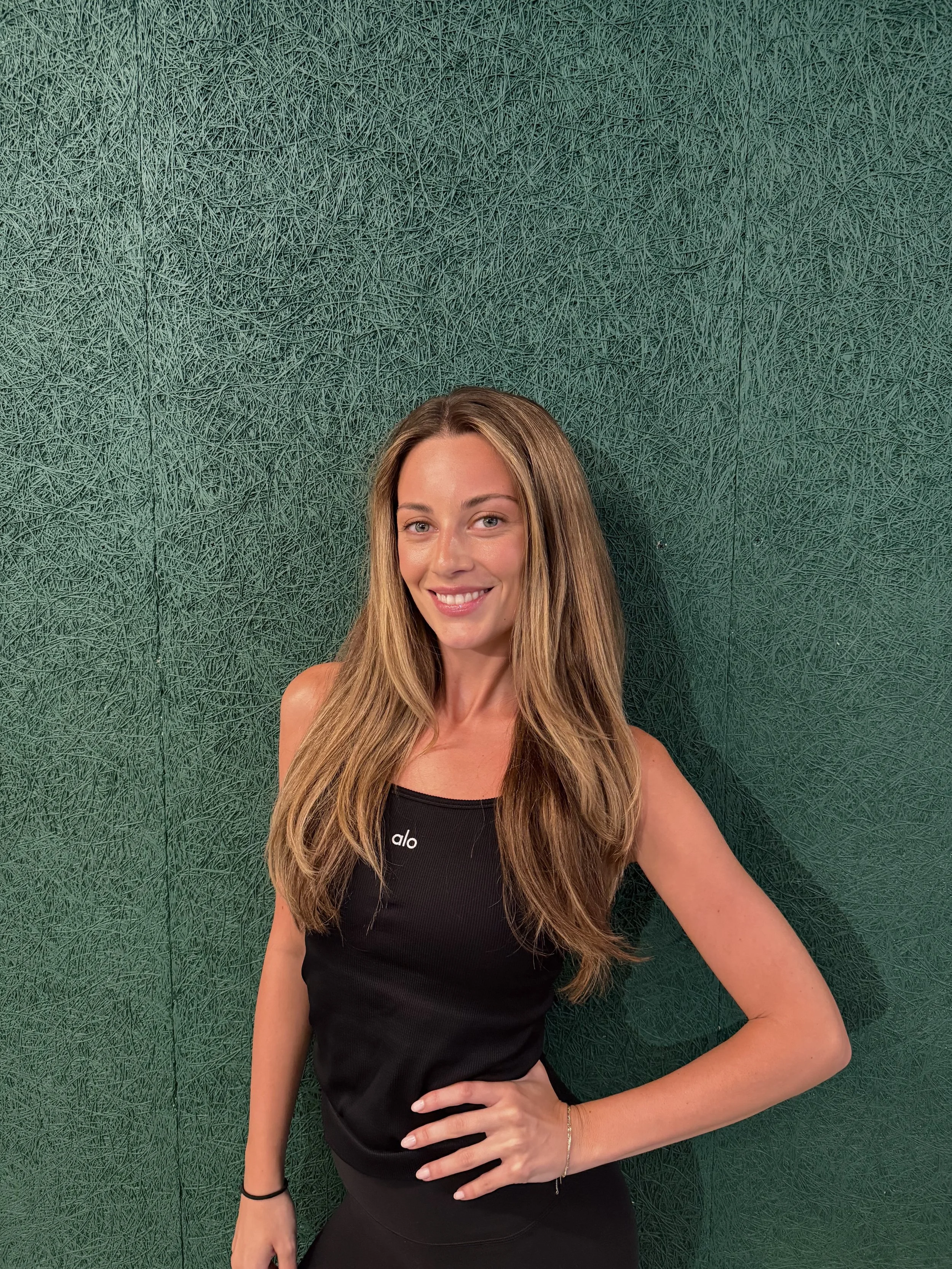 A woman with long, light brown hair and fair skin smiling, wearing a black sleeveless top, standing against a dark green textured wall.
