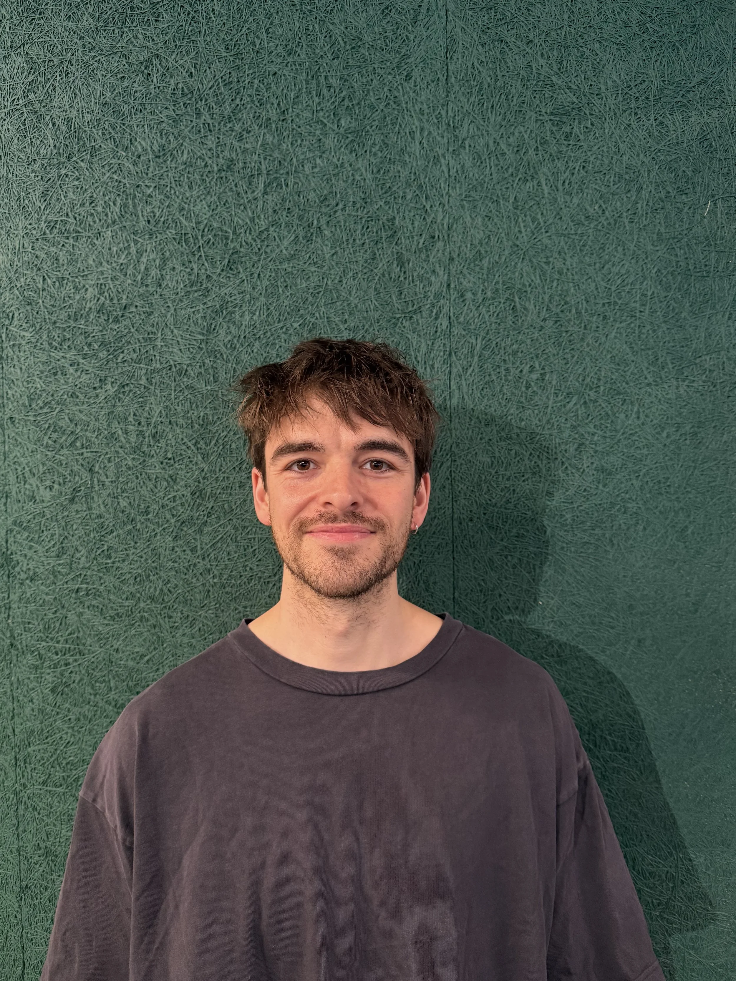 A young man with brown hair, light beard, wearing a black t-shirt, standing in front of a green textured wall, smiling.