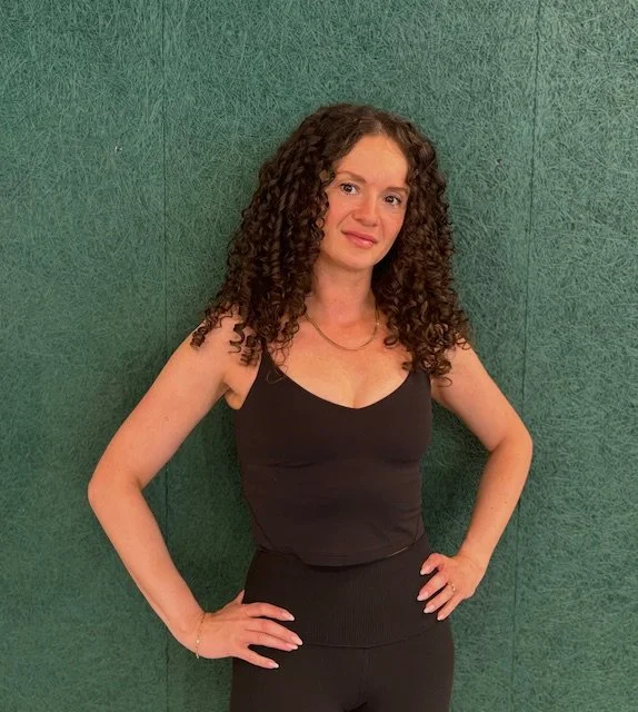 A woman with curly brown hair, wearing a black tank top and black pants, standing against a green textured wall.