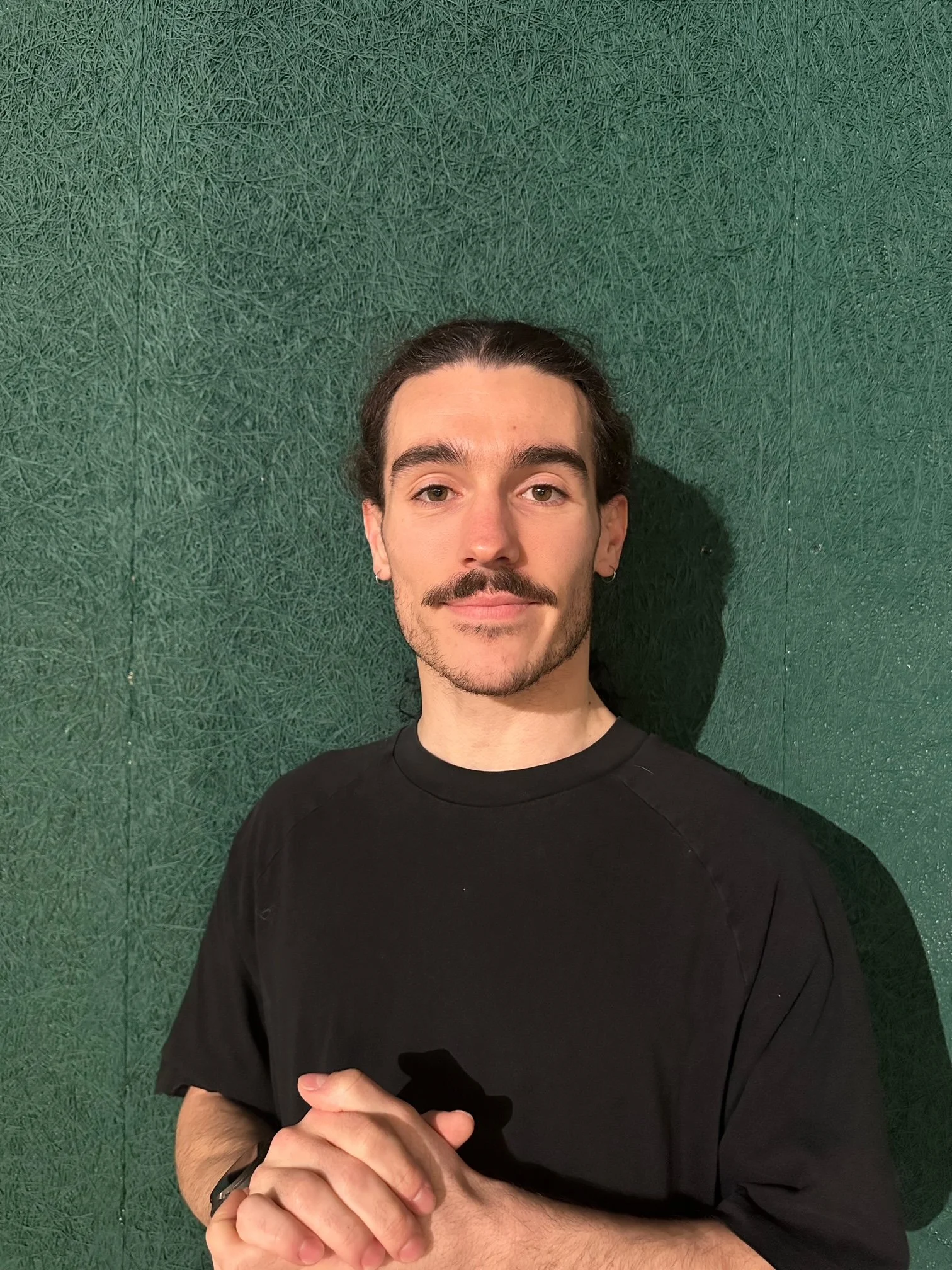A young man with long dark hair tied back, wearing a sleeveless black shirt and a black wristwatch, stands with arms crossed in front of a green textured background, smiling slightly.
