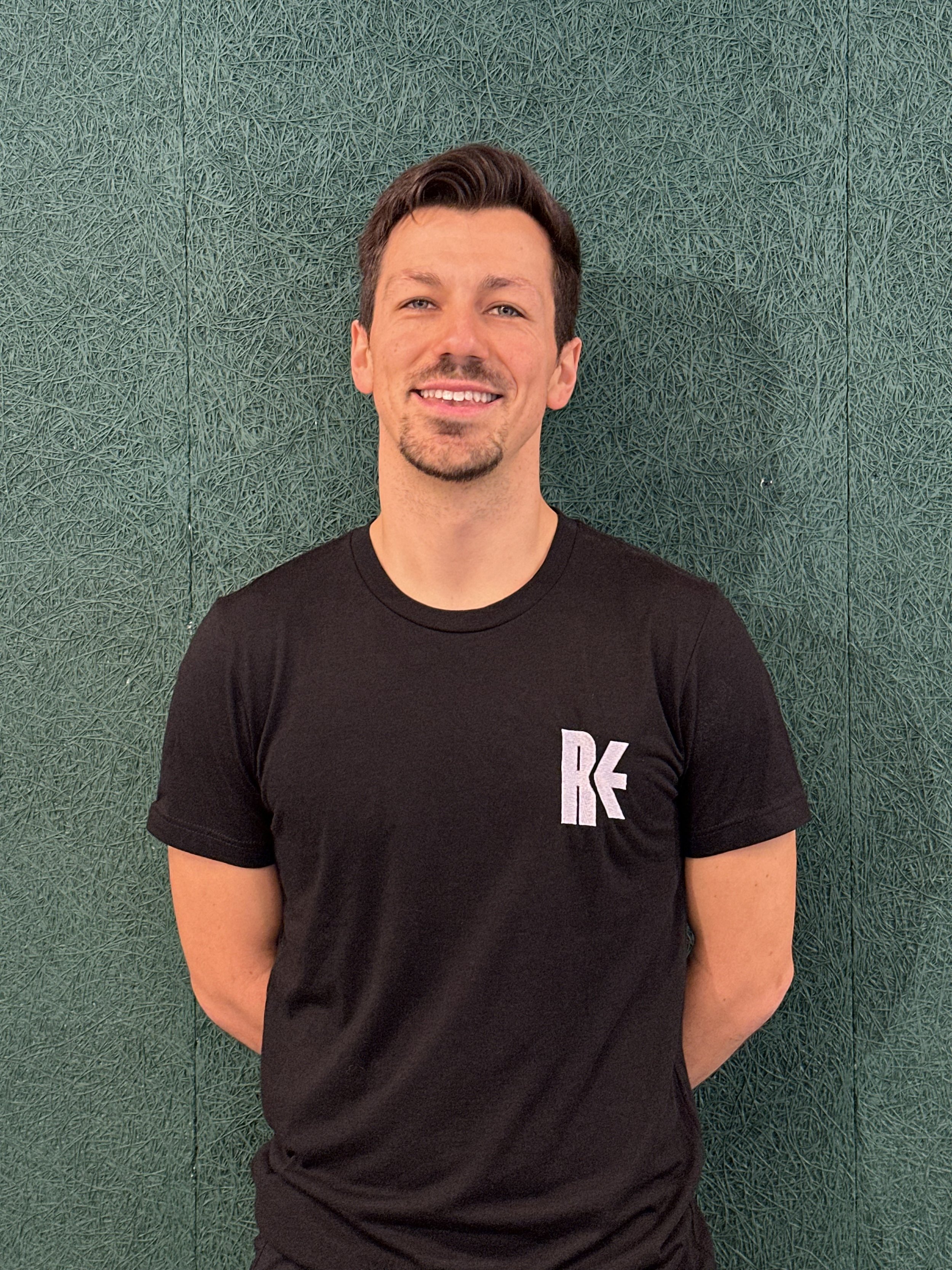 A smiling man with dark hair and a goatee, wearing a black T-shirt with a white logo, standing against a green textured wall.