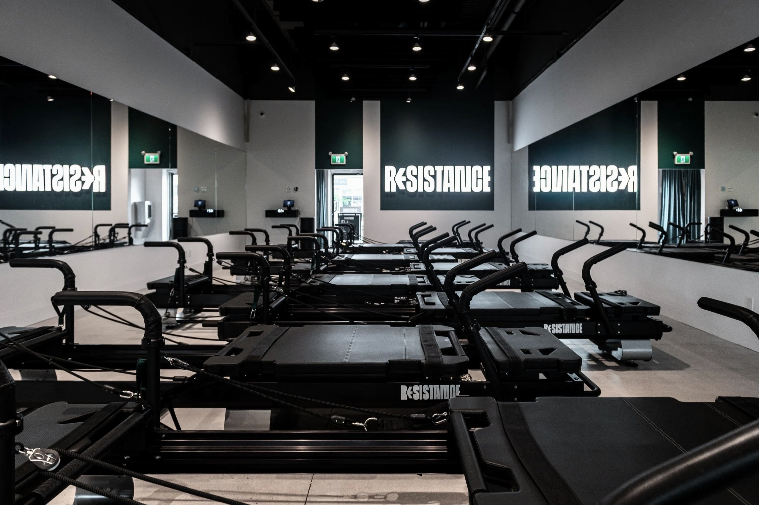 Empty fitness studio with multiple black resistance training machines arranged in rows, mirrors on the side walls, and a sign that reads 'RESISTANCE' on the back wall.