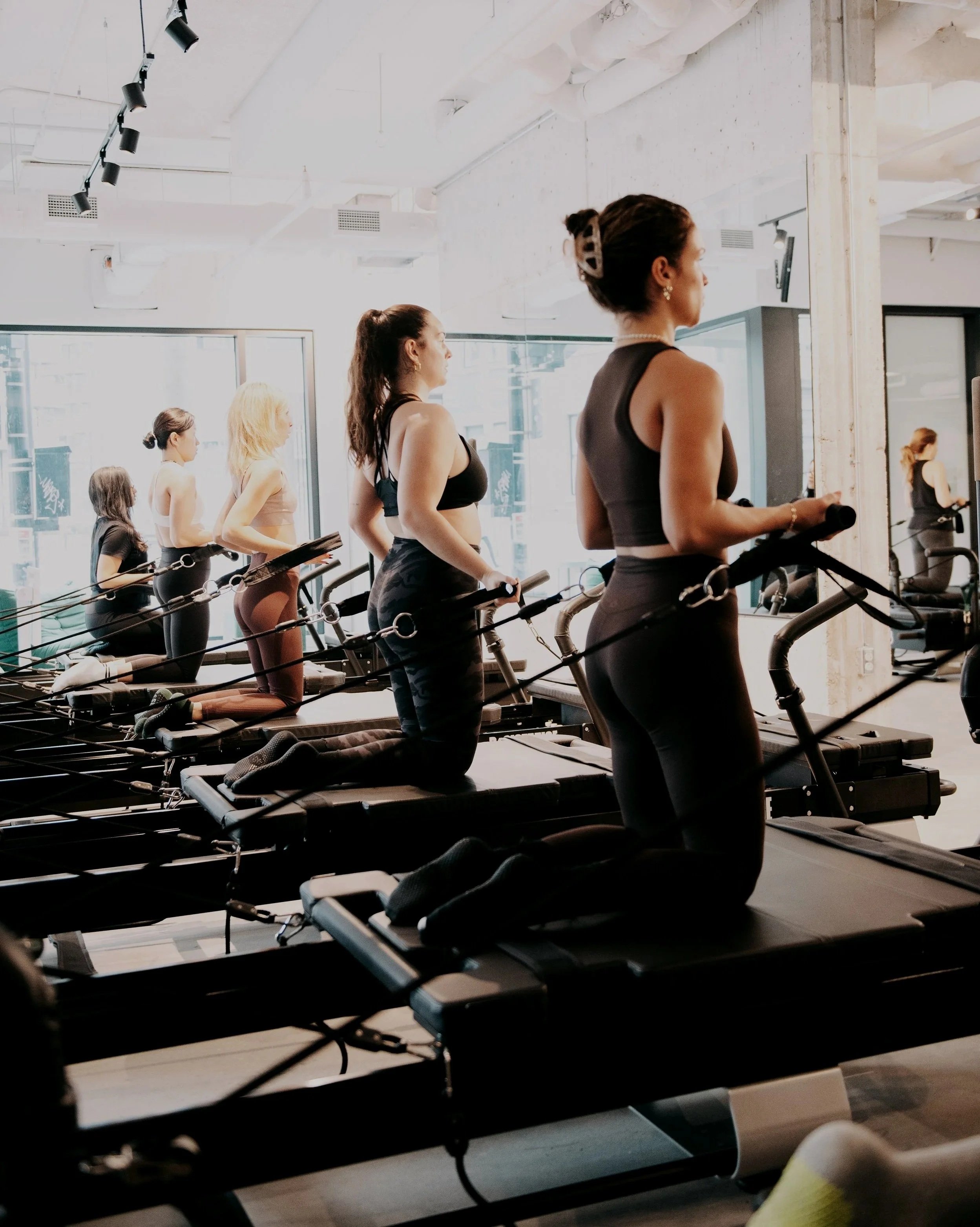 Women participating in a Pilates class on reformer machines in a modern, well-lit gym with large windows.