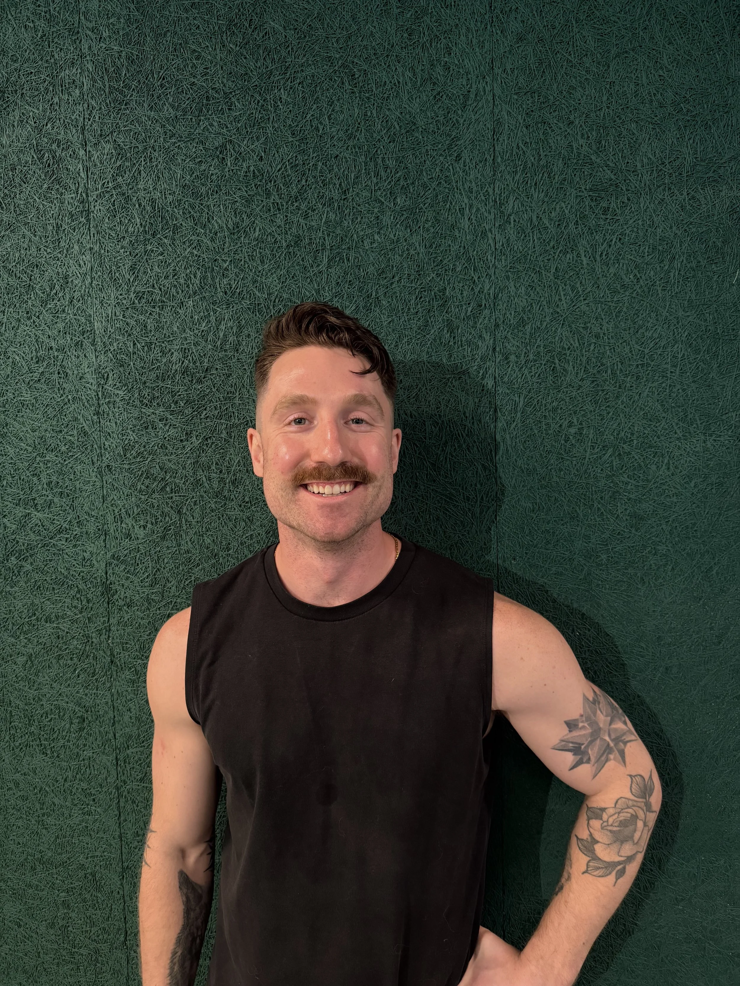 A smiling man with a mustache and tattoos on his arms, wearing a sleeveless black shirt, standing against a textured dark green wall.