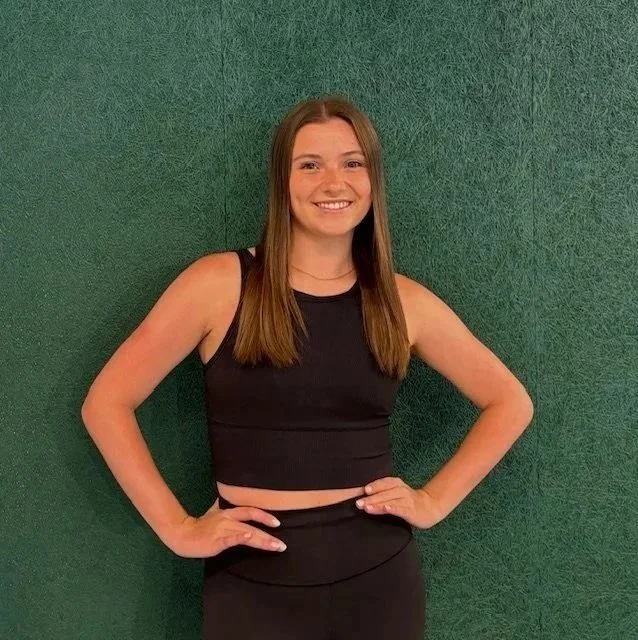A young woman with long brown hair smiling and posing with hands on hips in front of a green textured wall, wearing a black sleeveless top and black skirt.