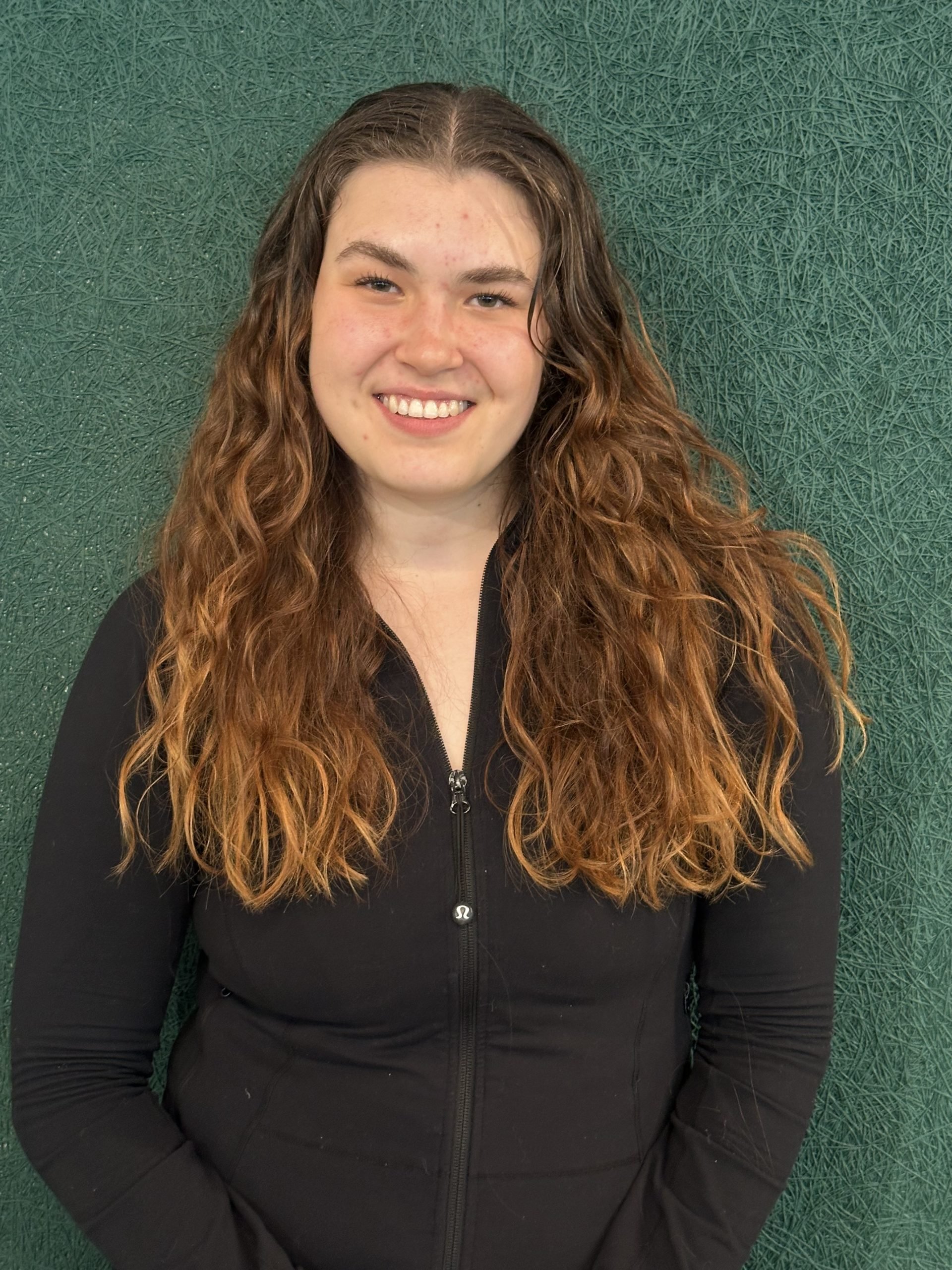 A young woman with curly auburn hair smiling and wearing a black zip-up jacket, standing against a textured green background.