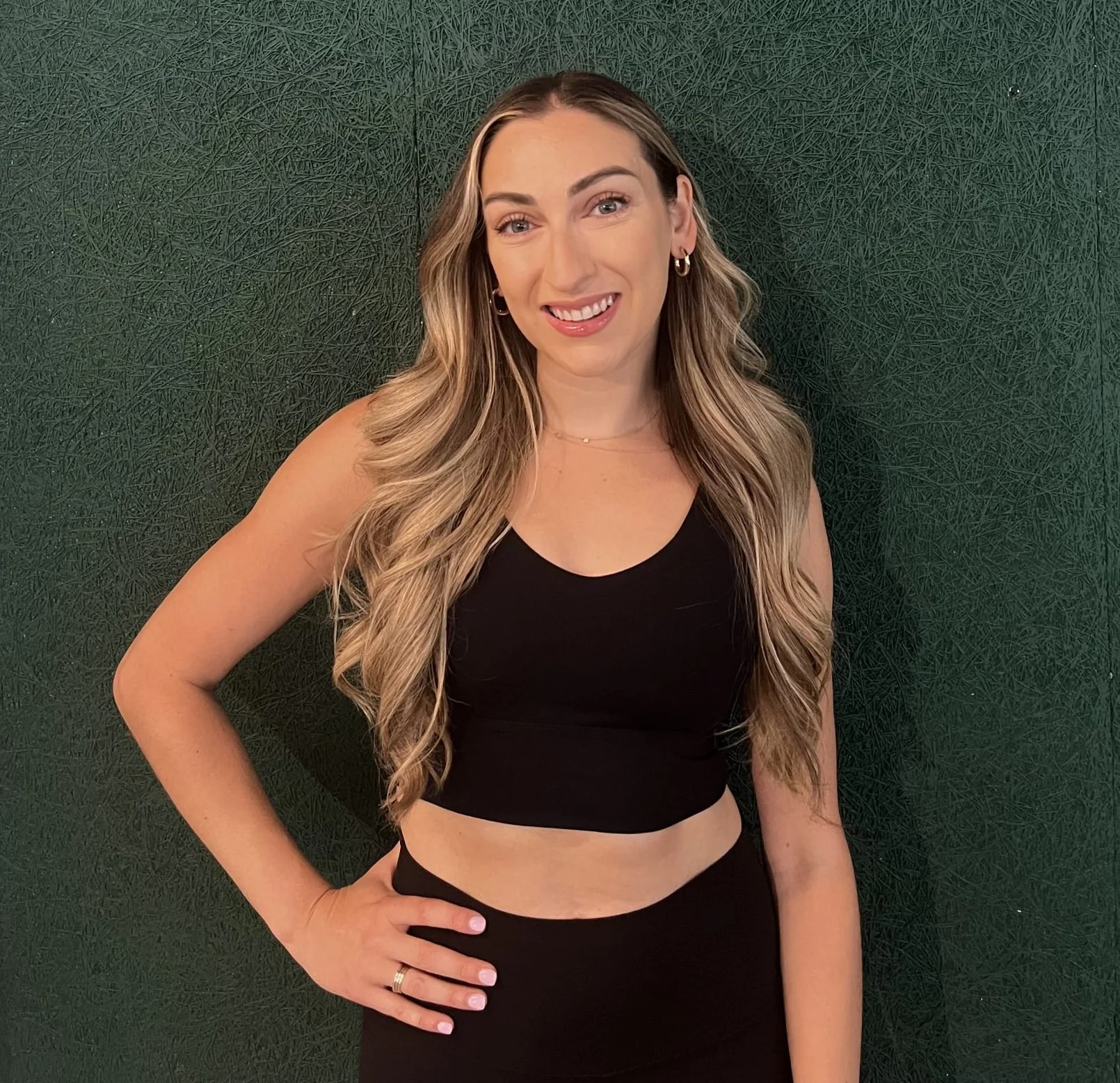 A woman with long, wavy blonde hair wearing a black sleeveless crop top and black pants, standing against a textured dark green background. She is smiling with her right hand on her hip.