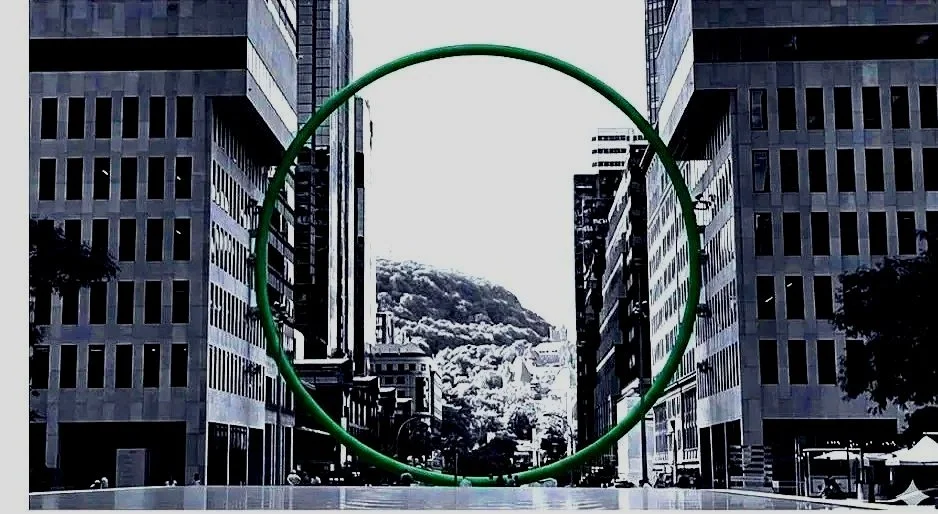 A large green circular sculpture in the middle of a city street between tall office buildings, with a mountain and cloudy sky in the background.