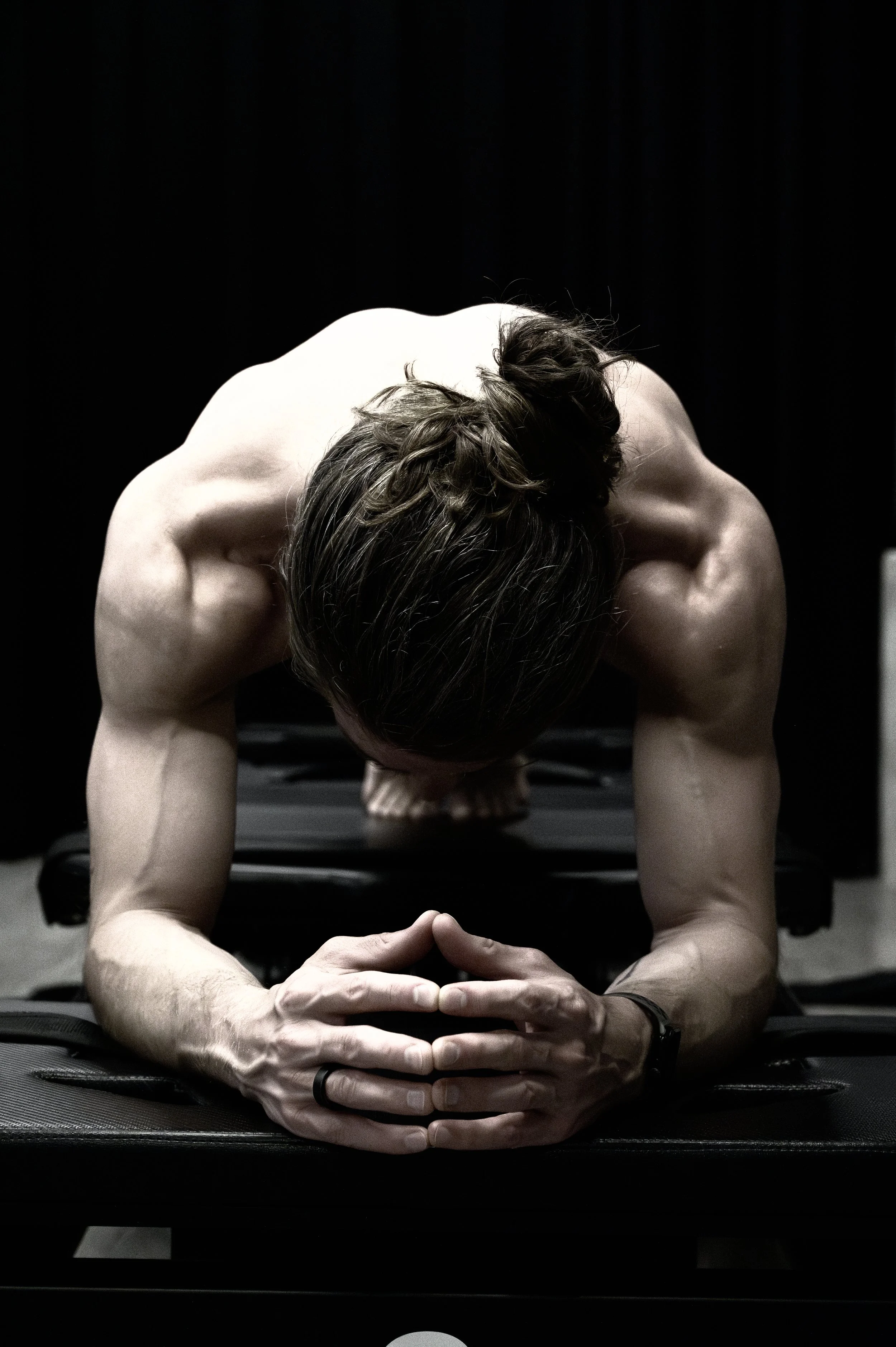 A shirtless man with long hair doing a plank exercise on a black mat in front of a black curtain.