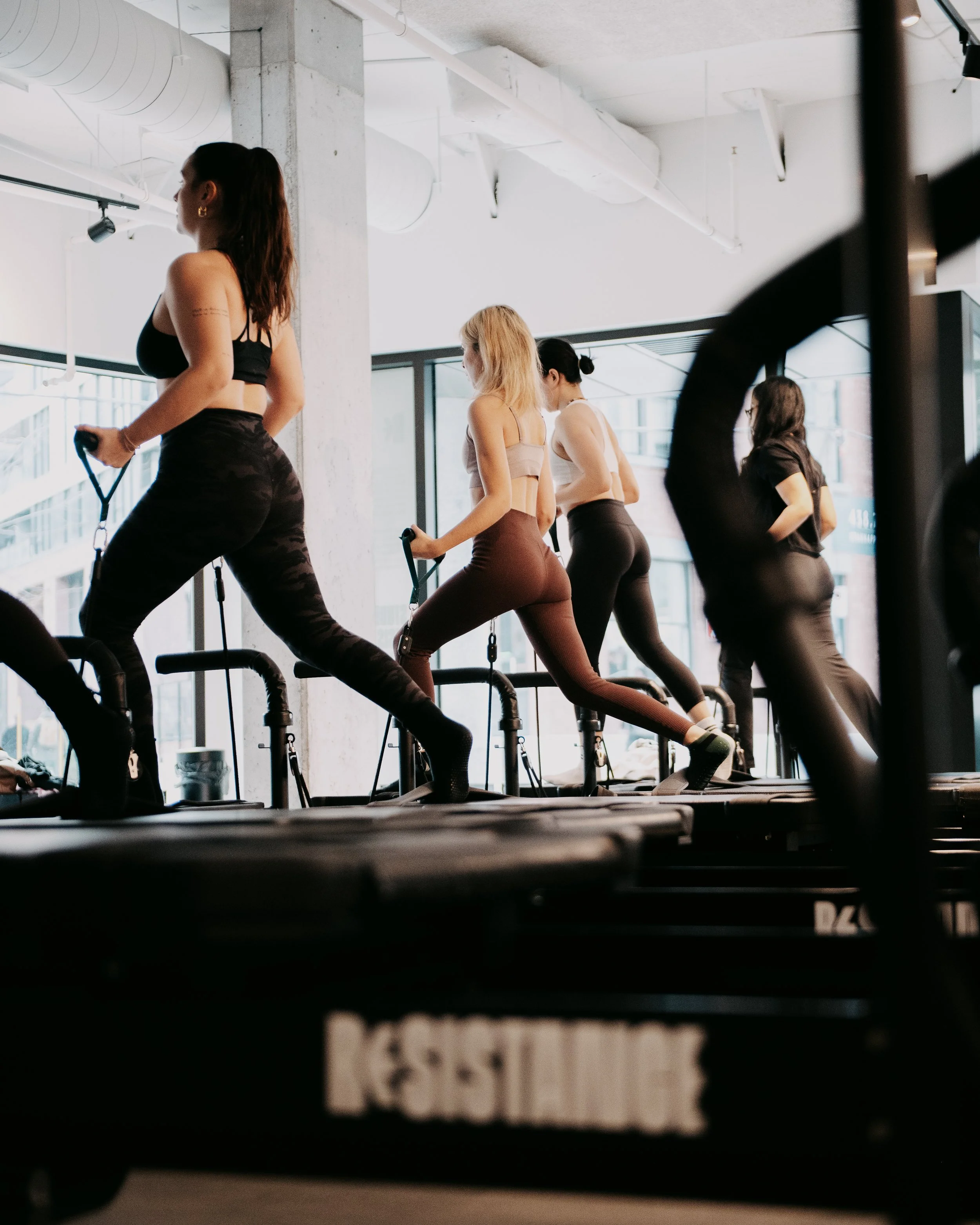 Four women participating in a fitness class on reformer machines in a modern gym with large windows and industrial design.