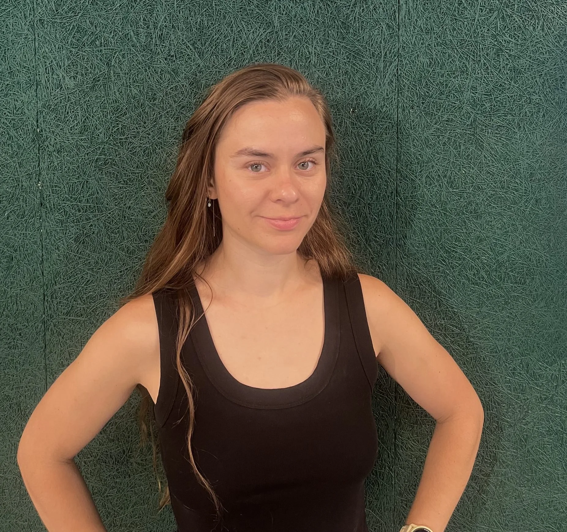A young woman with long, wavy brown hair wearing a black sleeveless top stands against a textured dark green background, smiling gently with her arms akimbo.