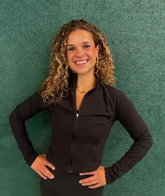 Young woman with curly hair smiling, wearing a black zip-up athletic jacket, standing against a textured green wall.