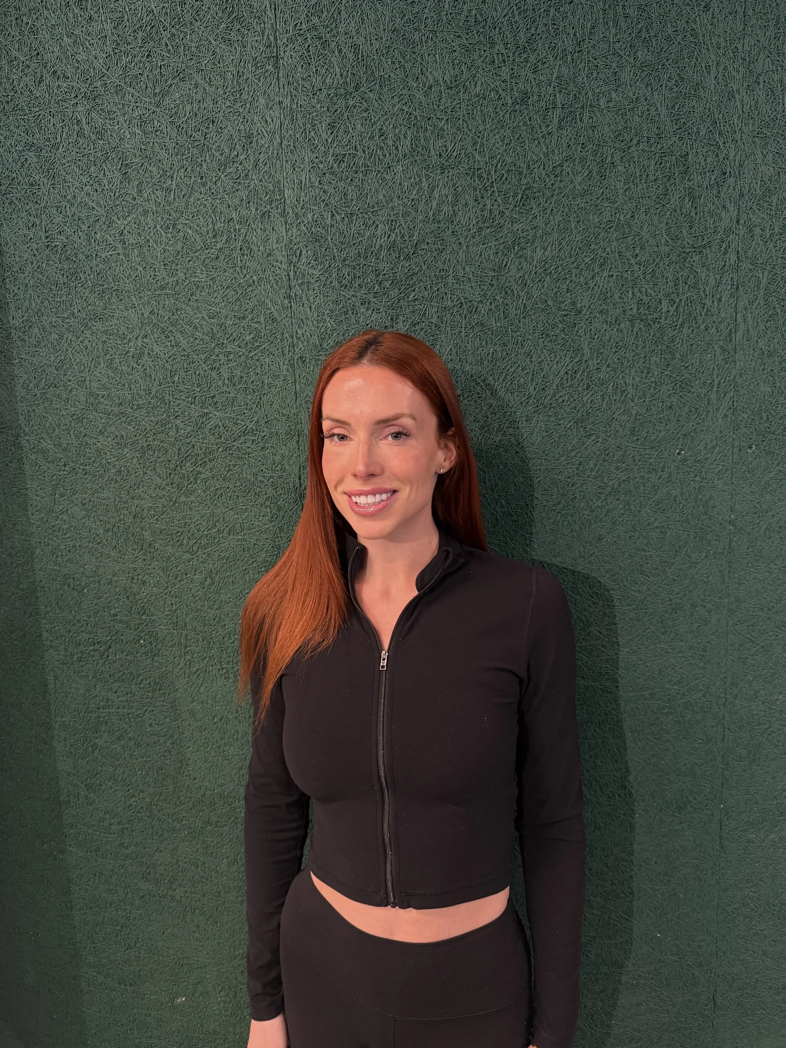 Woman with long red hair smiling, wearing a black zip-up jacket and black pants, standing against a green textured wall.