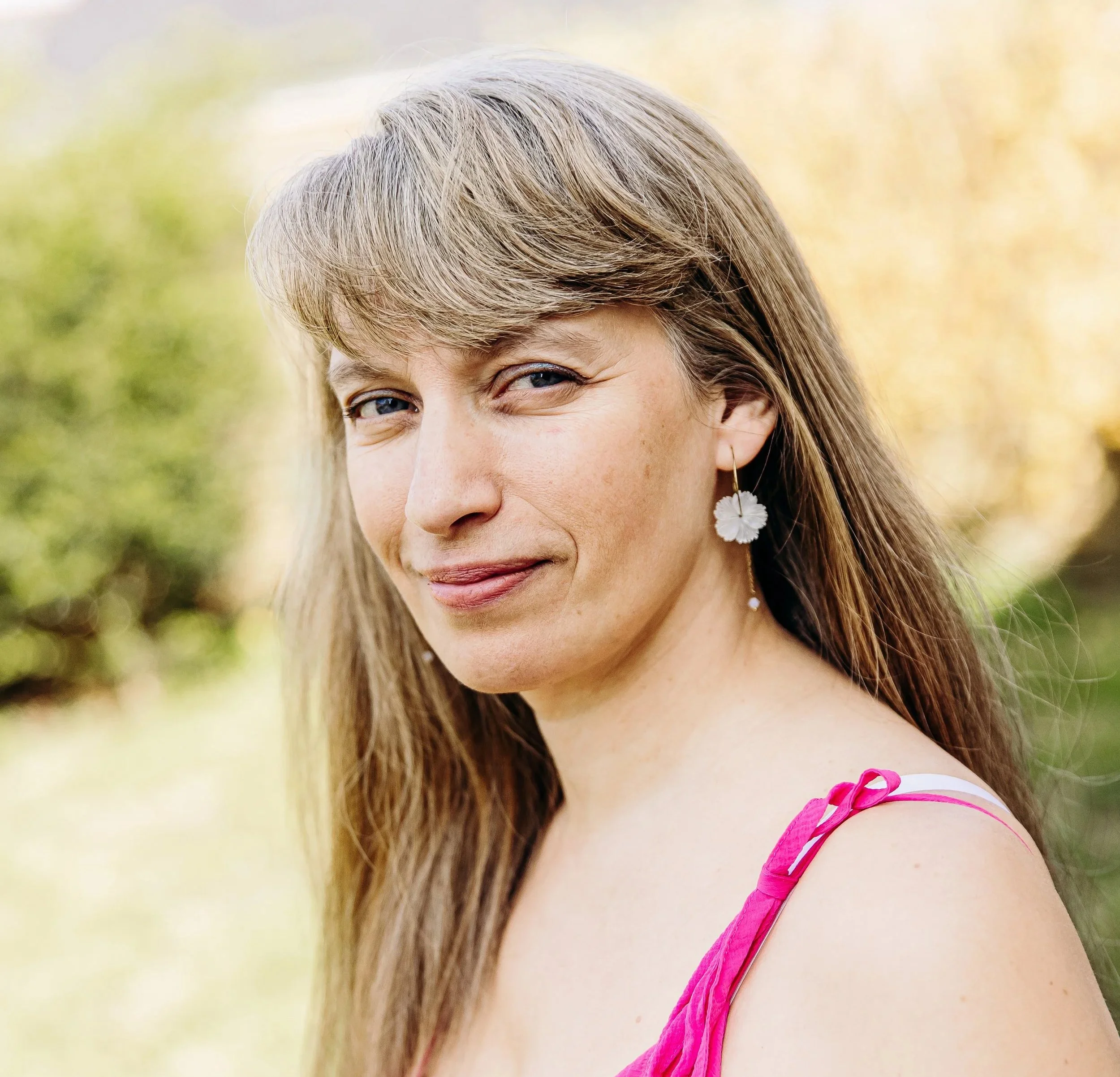 Un portrait partiel d'une femme avec des cheveux blonde, portant une boucle d'oreille en forme de fleur blanche et une robe rose, en extérieur avec un fond flou de nature verte.