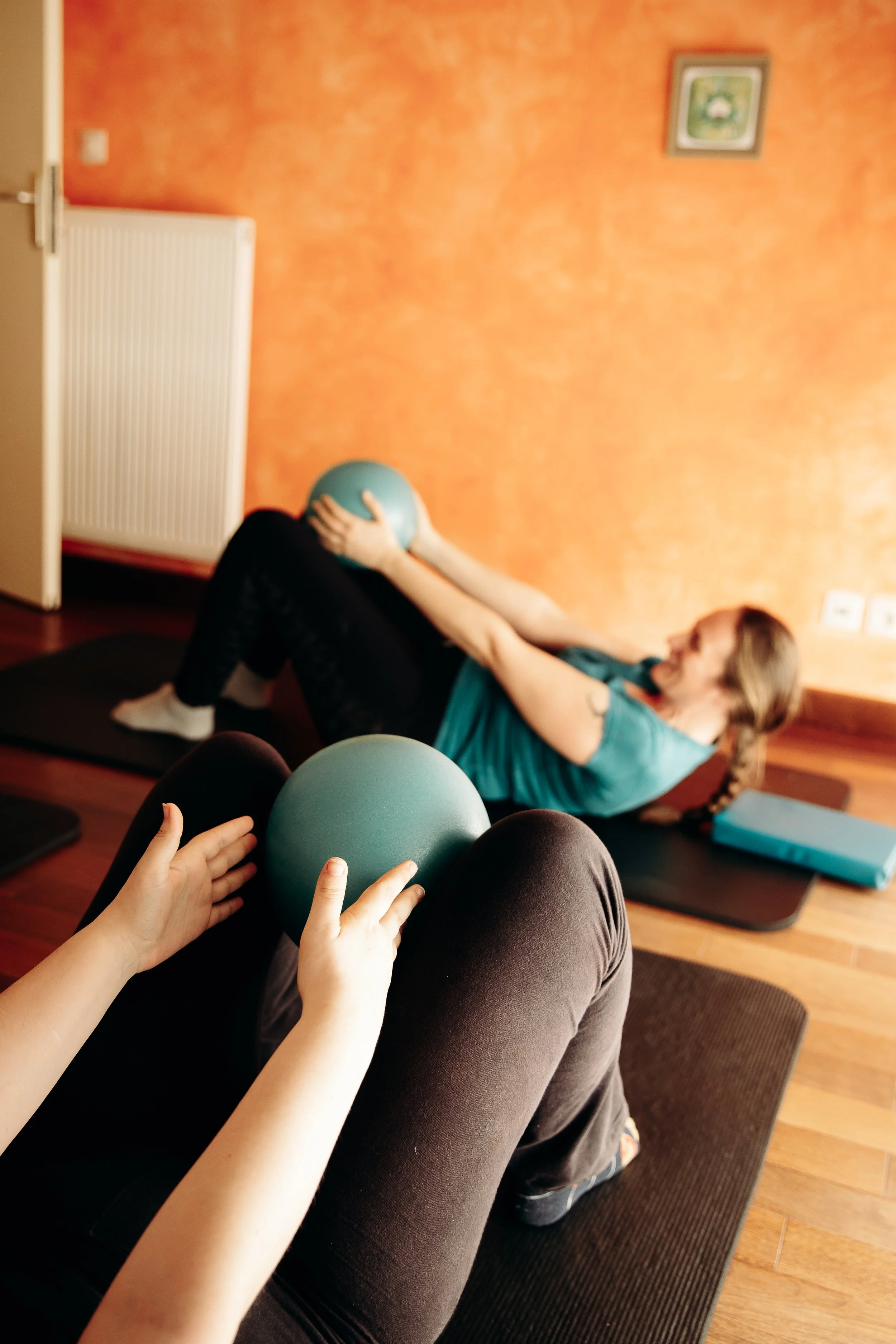 Femme faisant des exercices avec un ballon médecine dans une pièce orange, vue depuis les jambes d'une autre personne tenant un ballon.