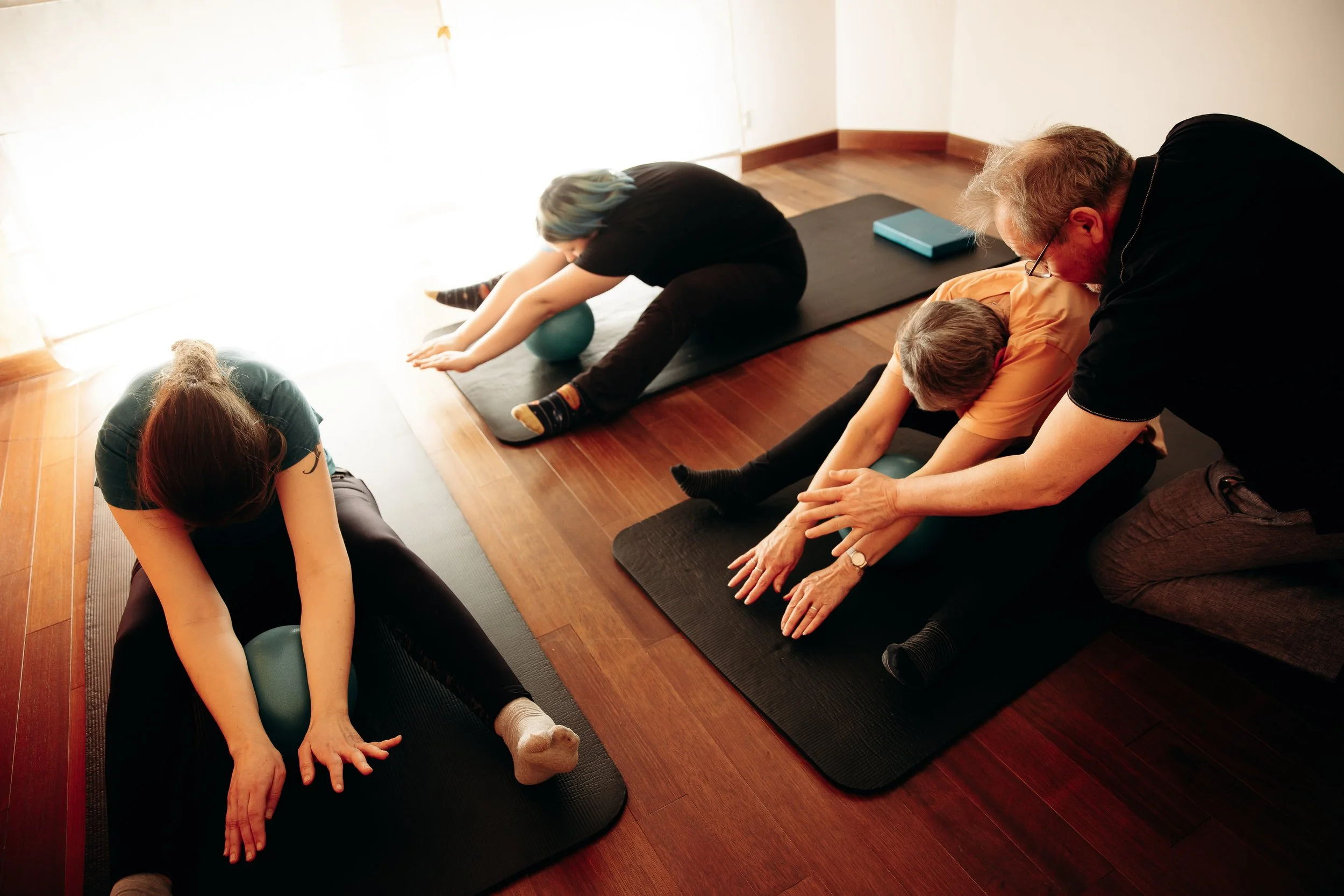 Groupe de quatre personnes faisant des étirements ou du yoga sur des tapis dans une pièce en bois, avec deux personnes aidant les autres.