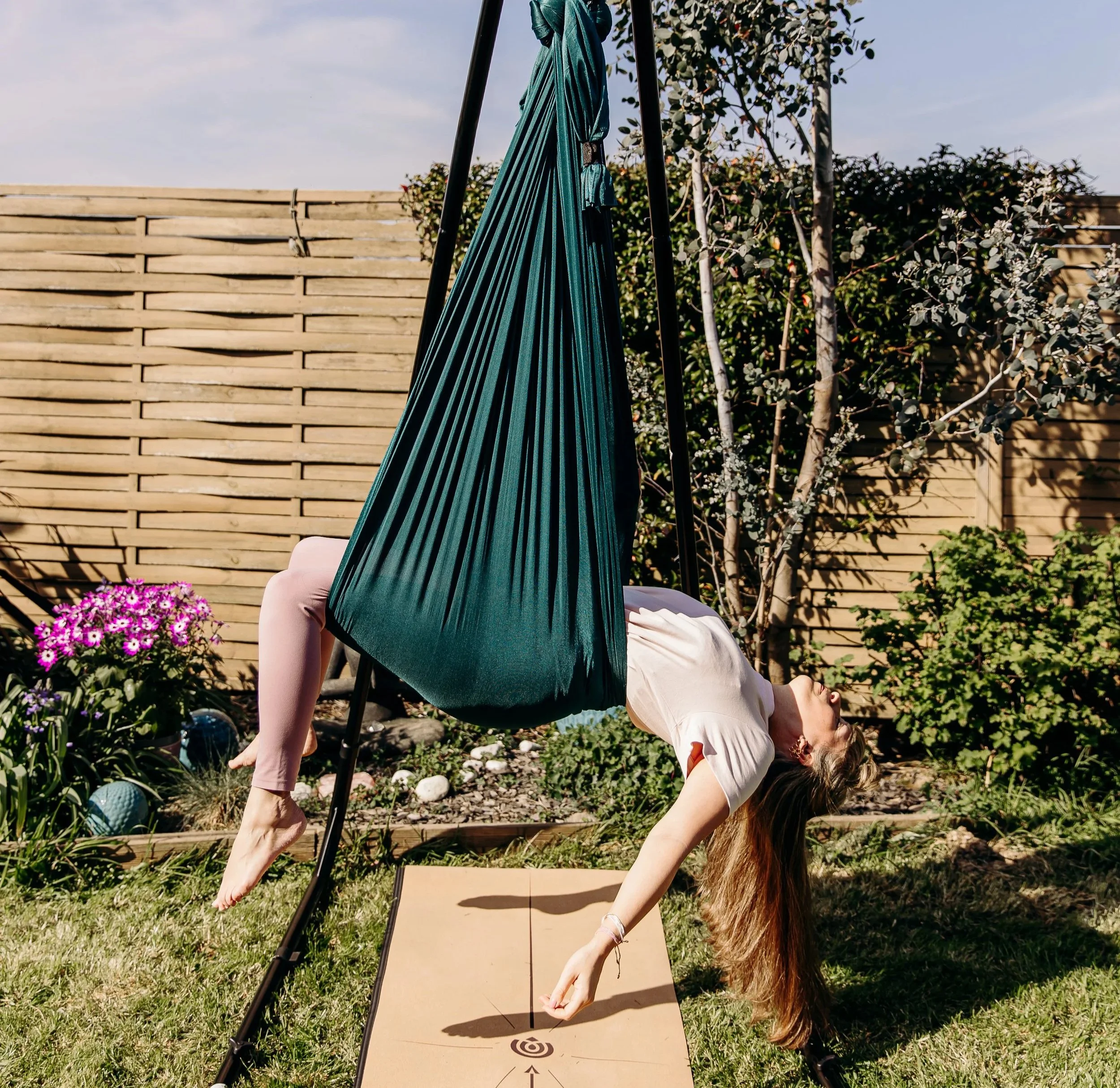 Une personne pratique le yoga en équilibre inversé sur une planche en extérieur, avec un parasol vert, une haie en arrière-plan, et des fleurs violettes à gauche.