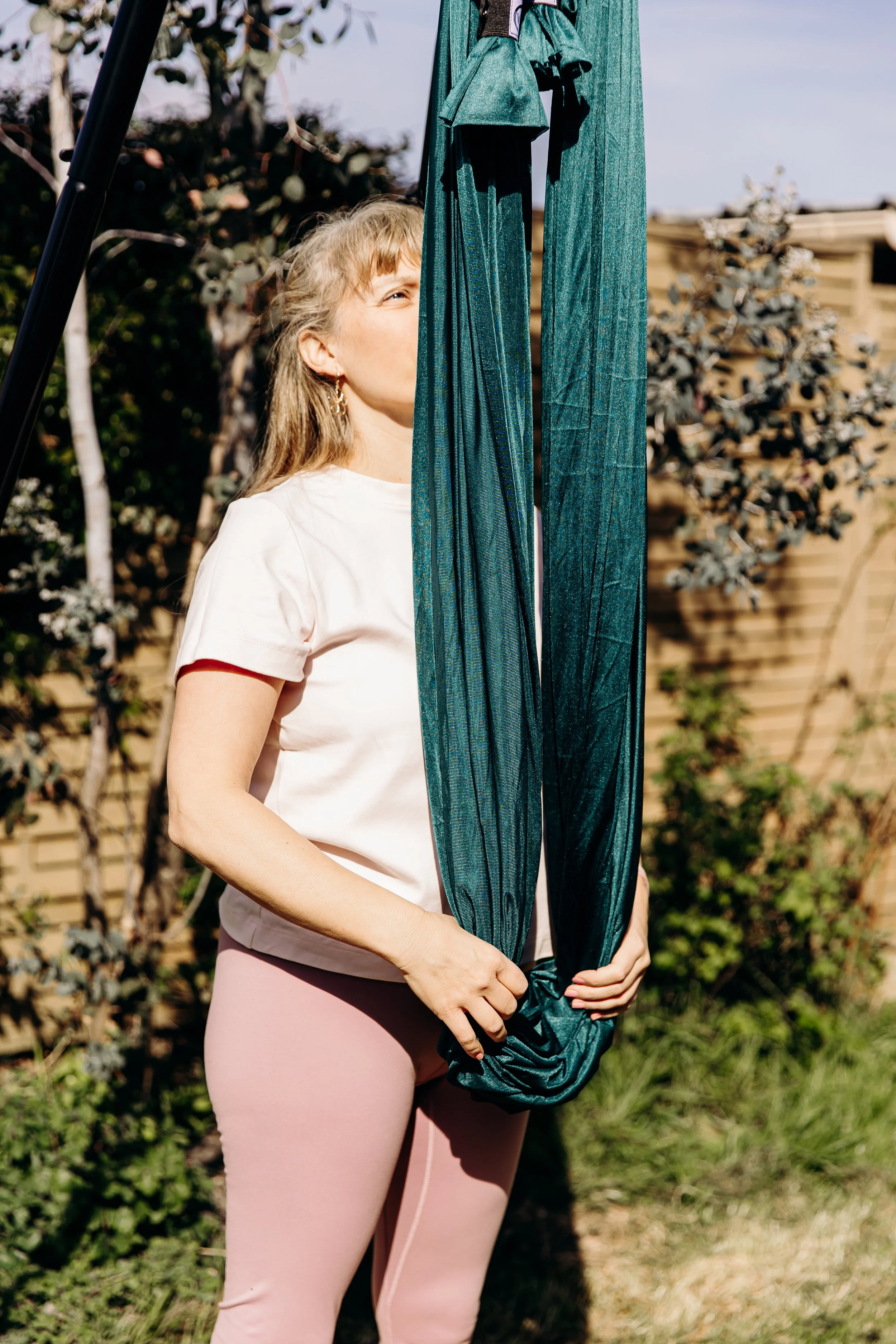 Une femme tenant un tissu bleu vert dans un jardin, avec un arbre et un terrain en arrière-plan.