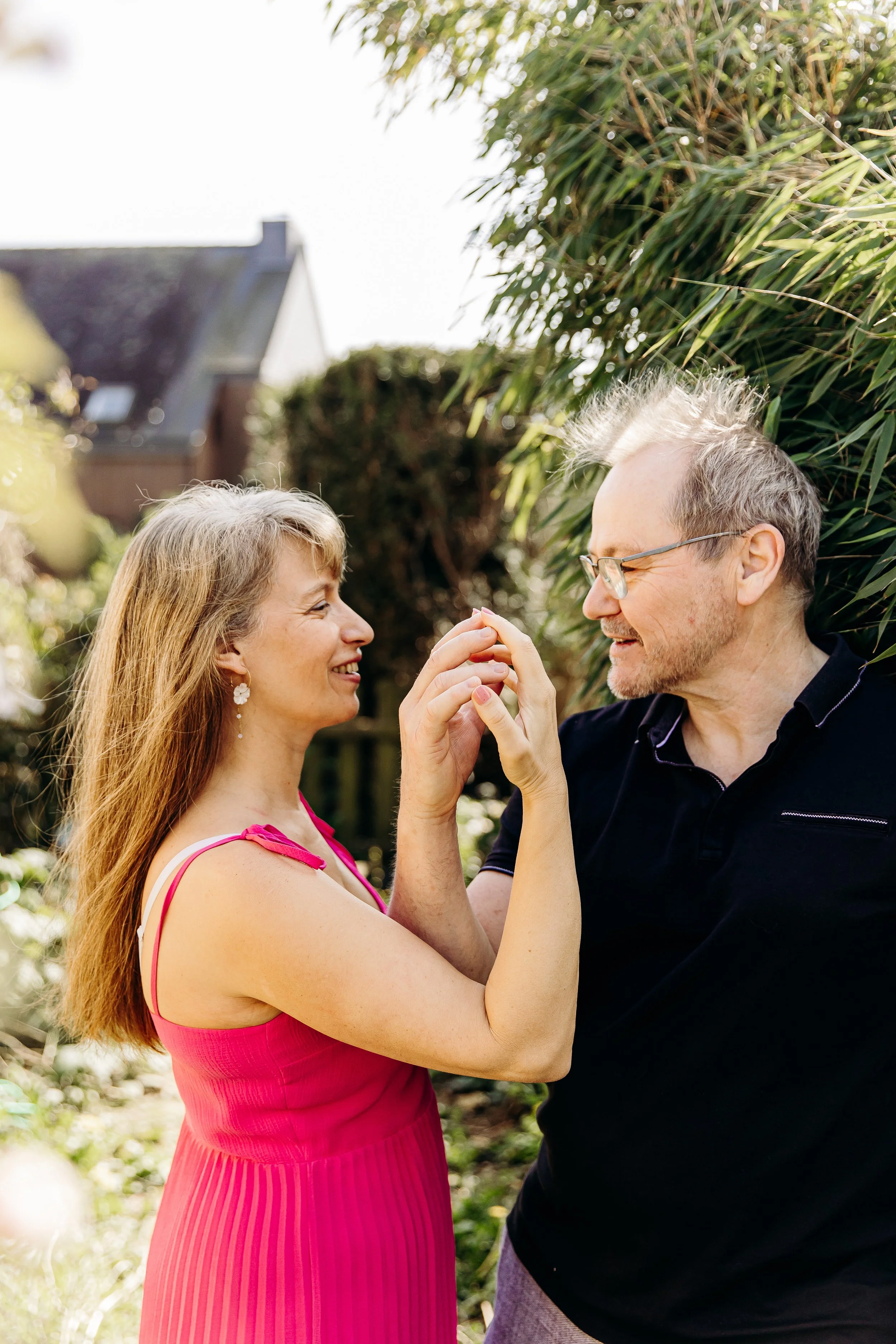 Un couple se tient dans un jardin, s'amusant et se frottant le nez, souriant, avec des arbustes et une maison en arrière-plan, sous une lumière naturelle.