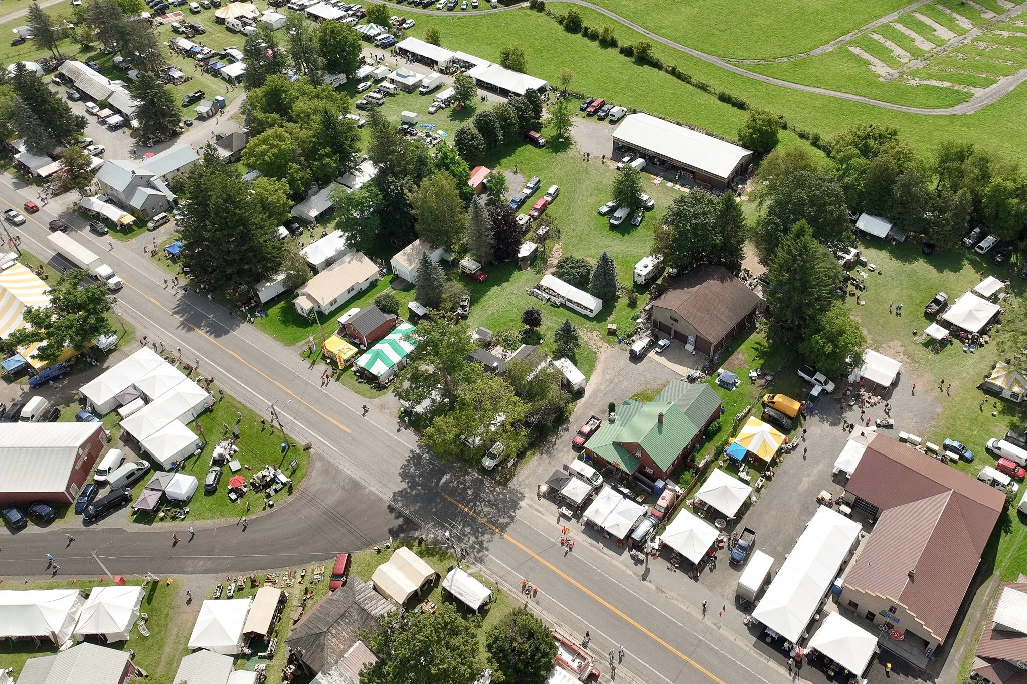 An aerial view of a small fair or market with tents, trees, and vehicles, situated along a main road through a town or village.