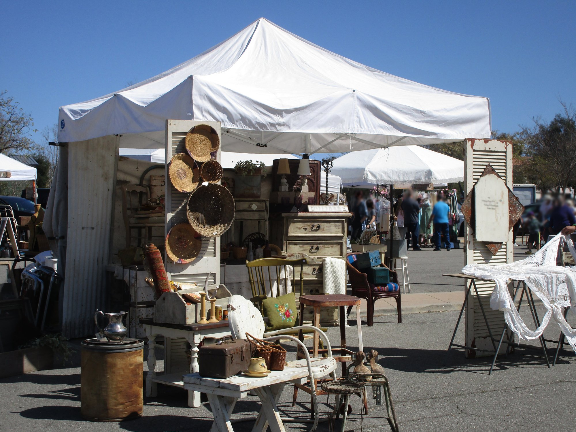 A white outdoor market tent with vintage home decor and furniture on display, including chairs, baskets, and decorative items.
