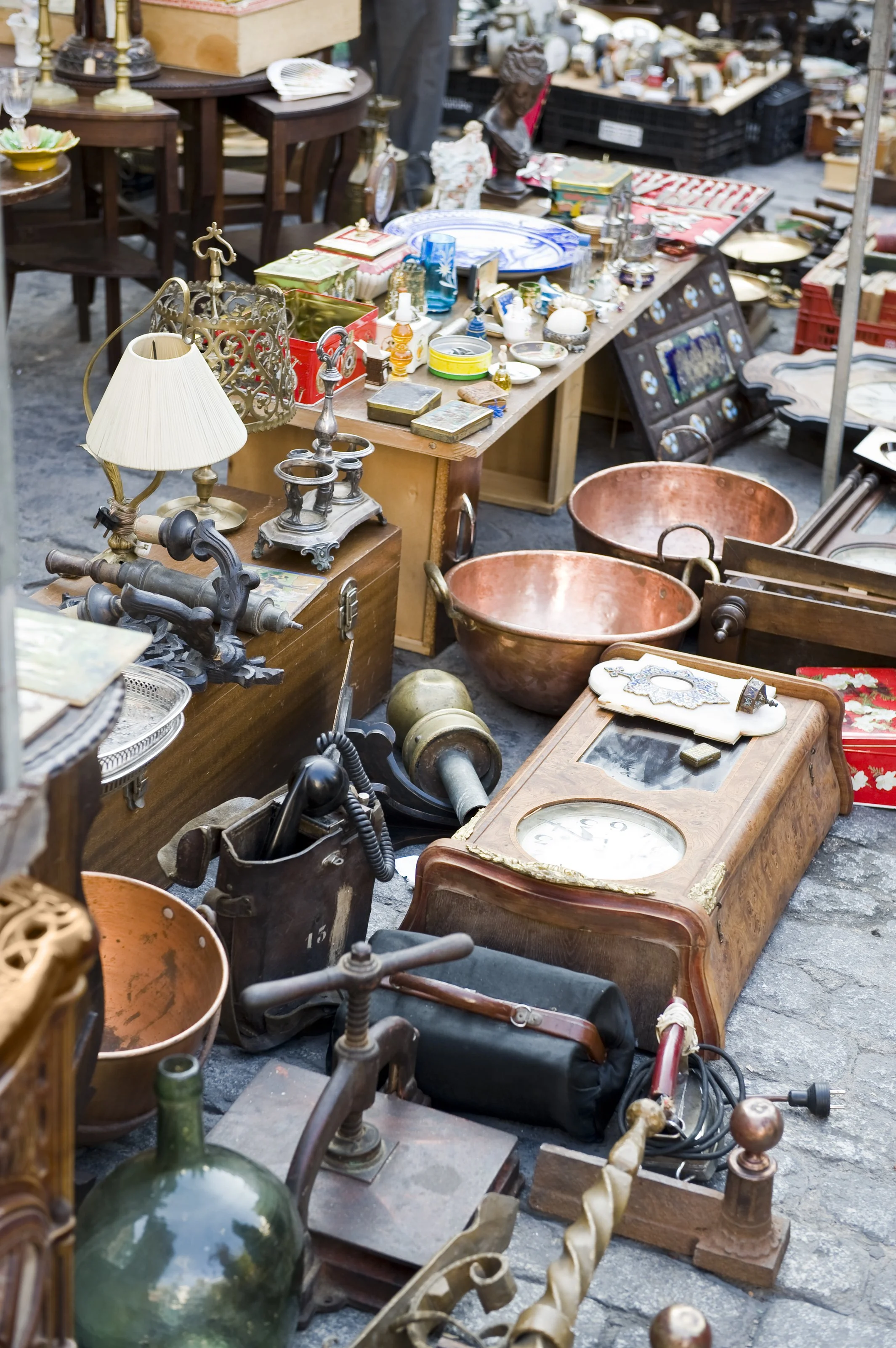 An outdoor flea market scene with various vintage and antique items arranged on tables and the ground, including bowls, lamps, clocks, and decorative objects.