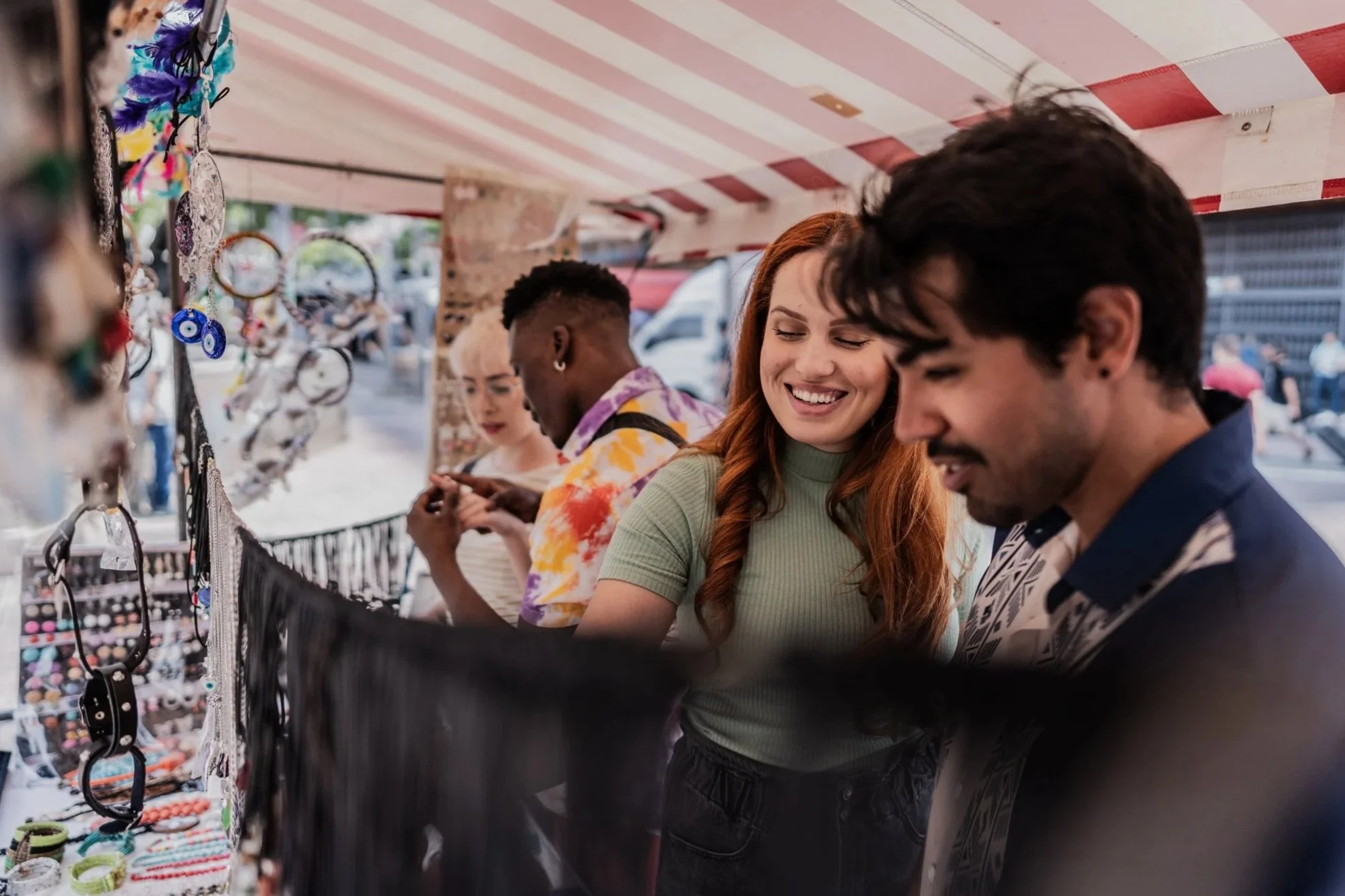 Two women and two men are shopping at an outdoor market stall with jewelry and accessories, smiling and browsing items.