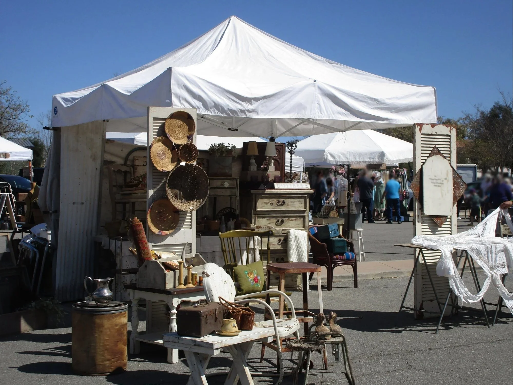 Outdoor flea market with various vintage items for sale under a white canopy, including baskets, furniture, and decorative objects, with people browsing in the background on a sunny day.