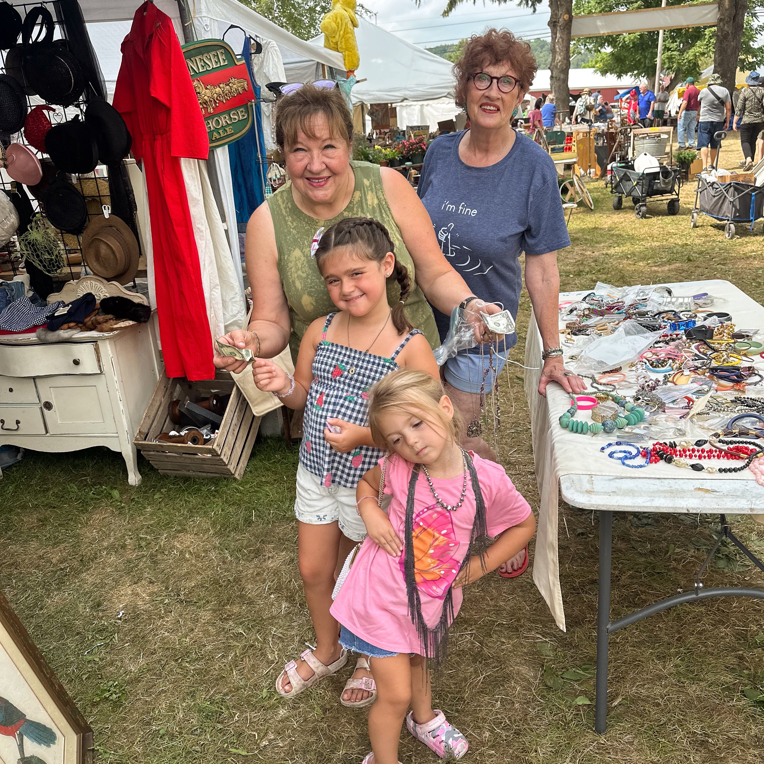 Three children and two women at an outdoor craft fair, with tables displaying jewelry and accessories in the background.