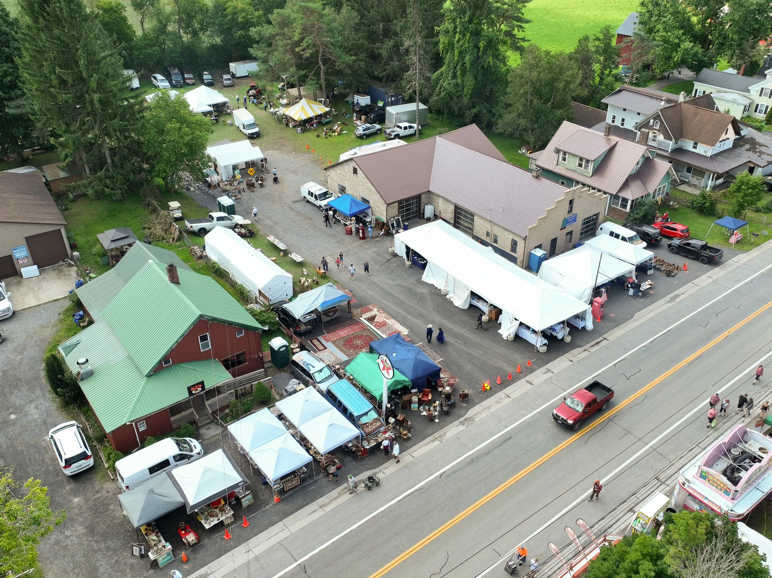 Aerial view of an outdoor market with white tents set up along a street, with people walking and shopping, surrounded by houses and trees.