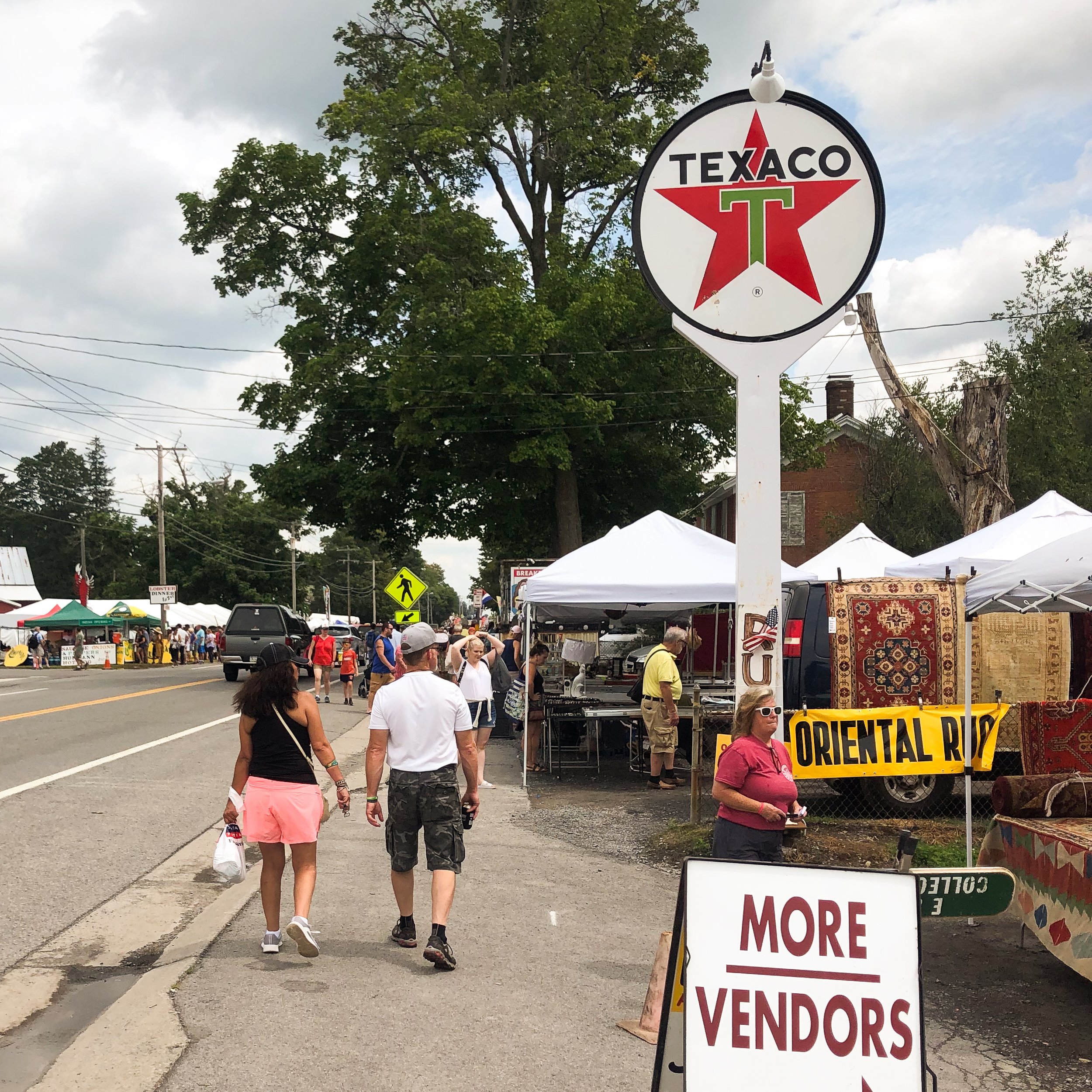 People walking past vendor tents on a street during a fair or market event with a large Texaco sign overhead and a sign reading "More Vendors" in front of the tents.