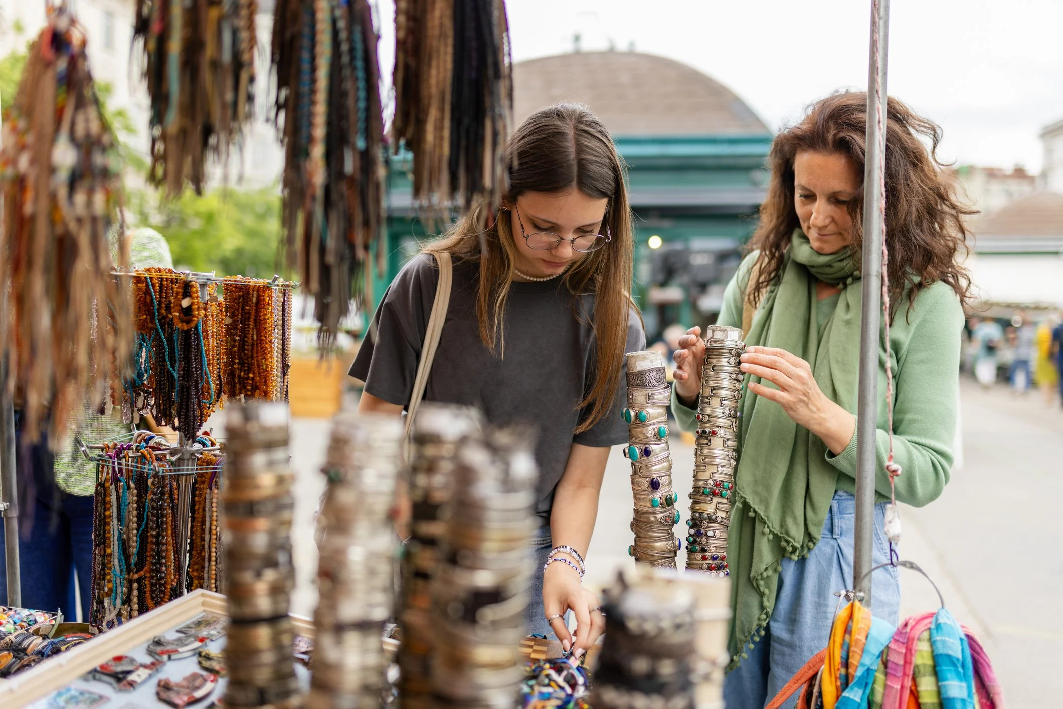 Two women browsing jewelry at an outdoor market stall. The stall displays bracelets, necklaces, and other accessories. The background shows a building and other market activity.