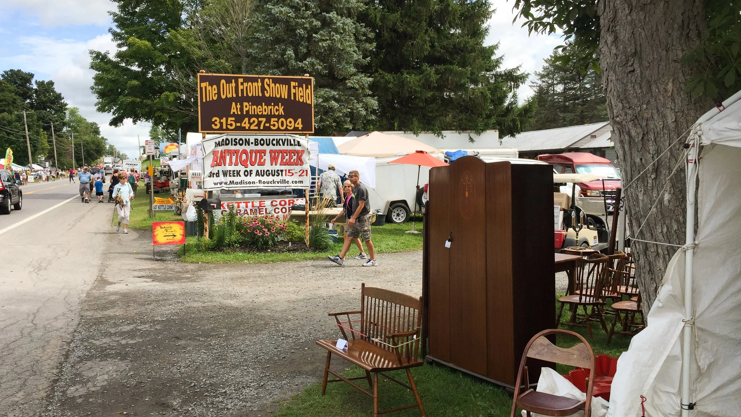 A busy street market with tents and stalls, a sign advertising antique week, and people walking and shopping during daytime.