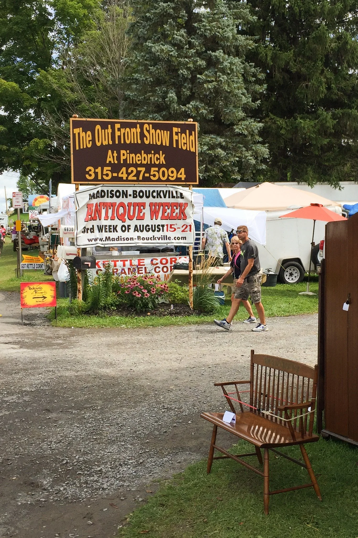 People walking past a sign at an outdoor antique market during Madison-Bouckville Antique Week, held from August 15 to 21, with vendor tents and trees in the background.