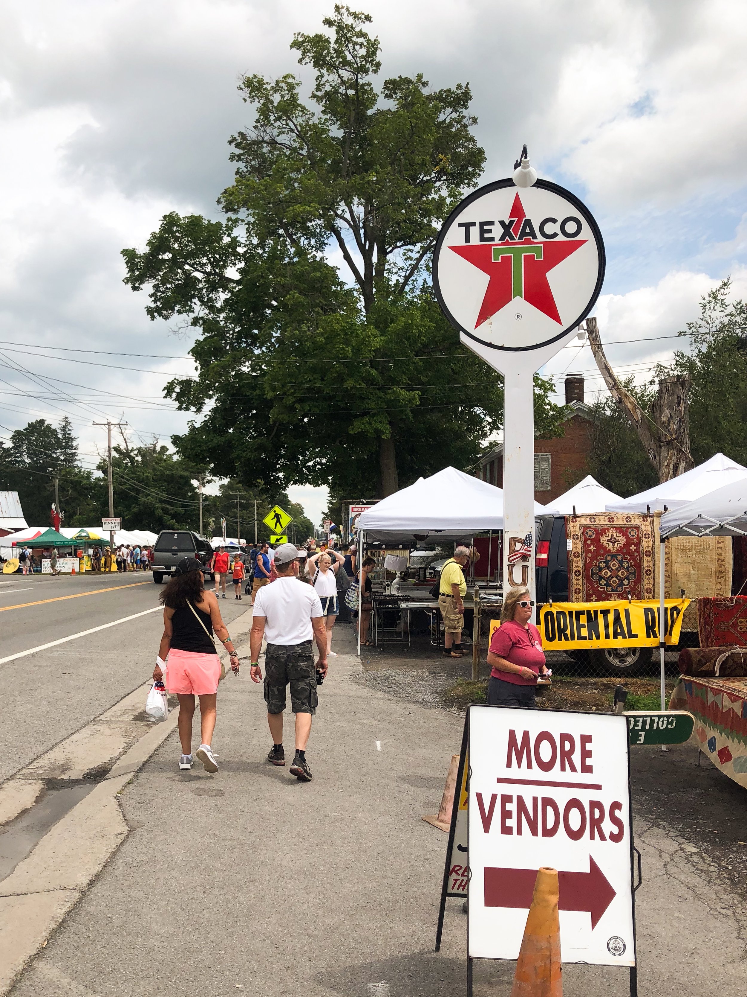 Street fair with vendor tents, a sign for more vendors pointing right, people walking on sidewalk, and a Texaco sign.
